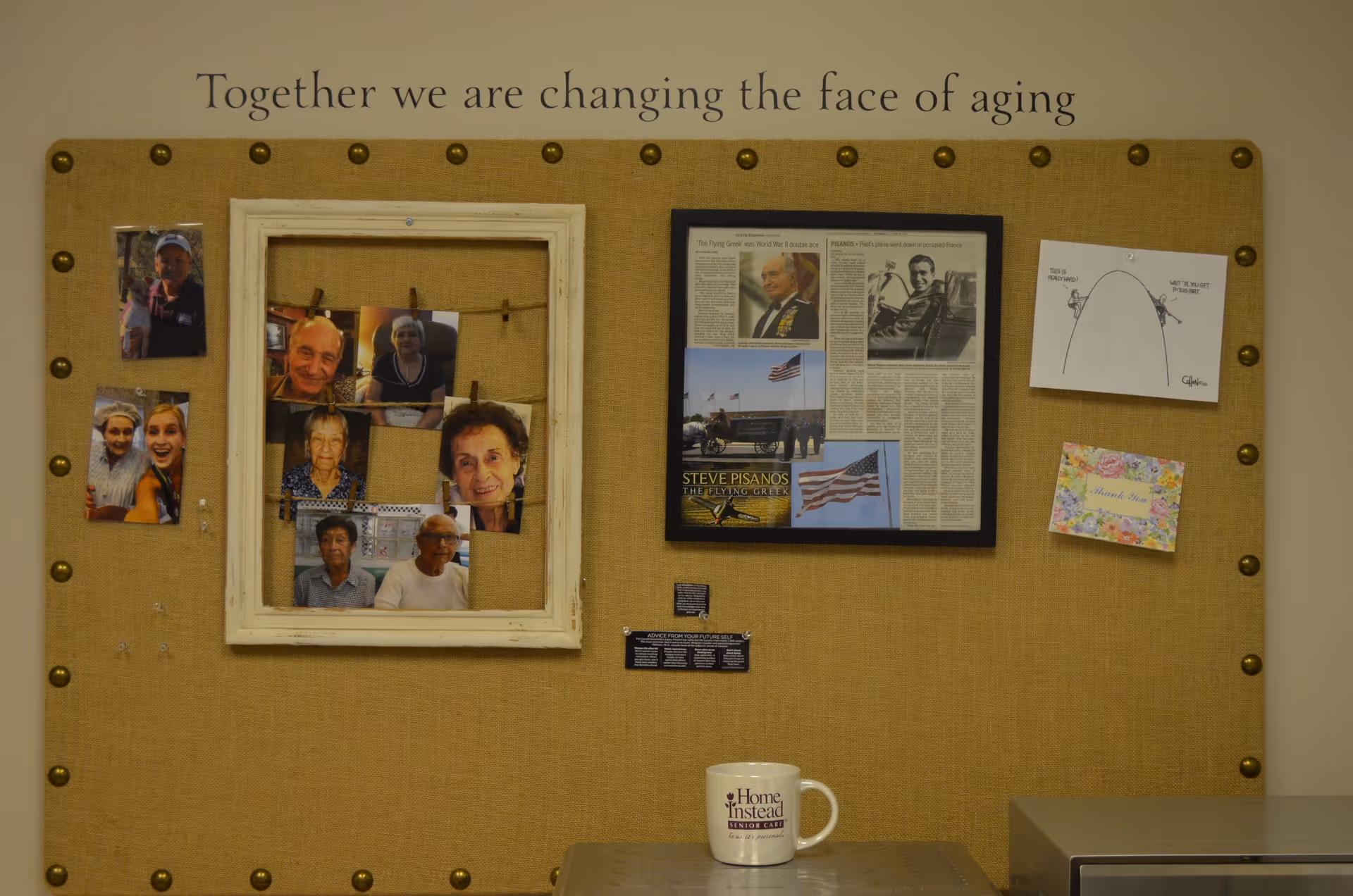 Beige bulletin board displays photos, articles, and a Home Instead mug on a nearby table.