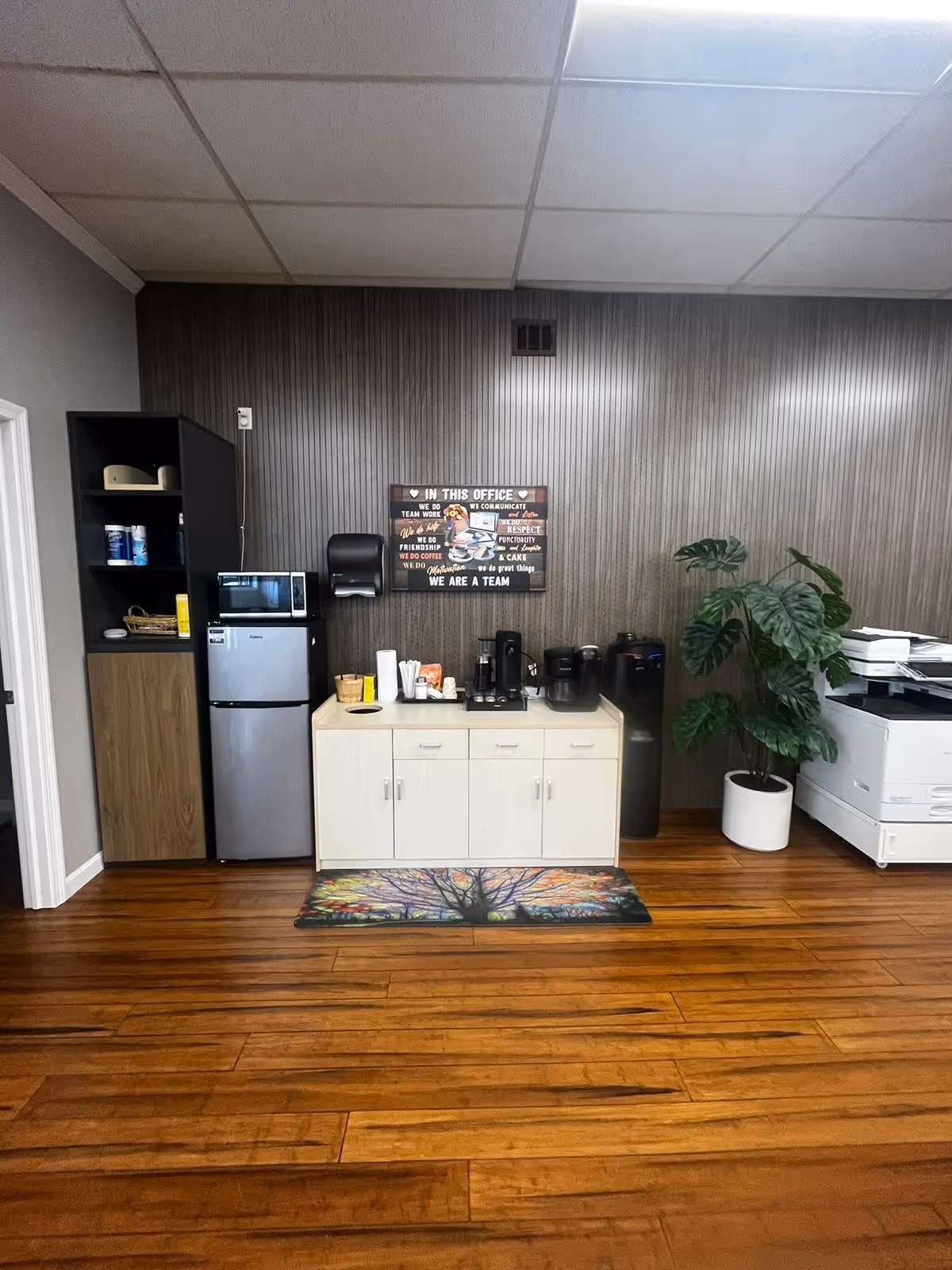 Office kitchenette with coffee station, small fridge, microwave, cabinets, wall sign, and a potted plant.