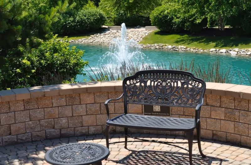 An ornate metal bench and round table sit beside a turquoise pond with a fountain.