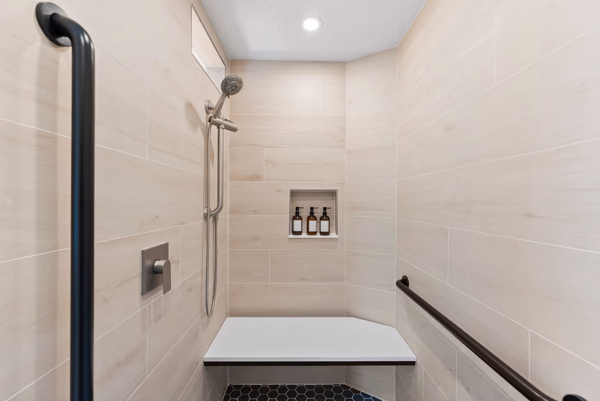 Accessible shower with beige tiled walls, a bench, handheld shower, and built-in shelf.