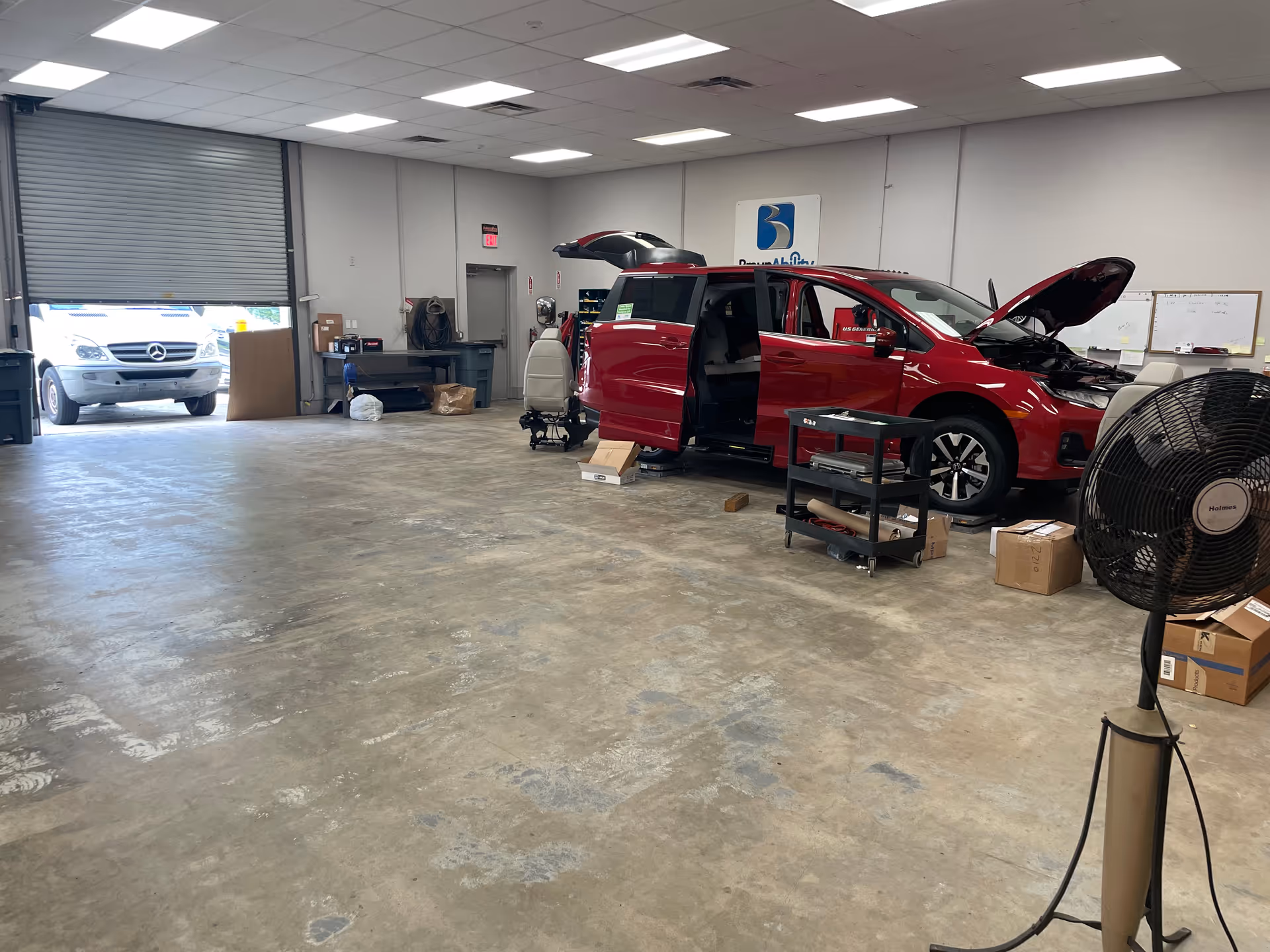Red minivan being serviced inside a large workshop with tools, a fan, and boxes.