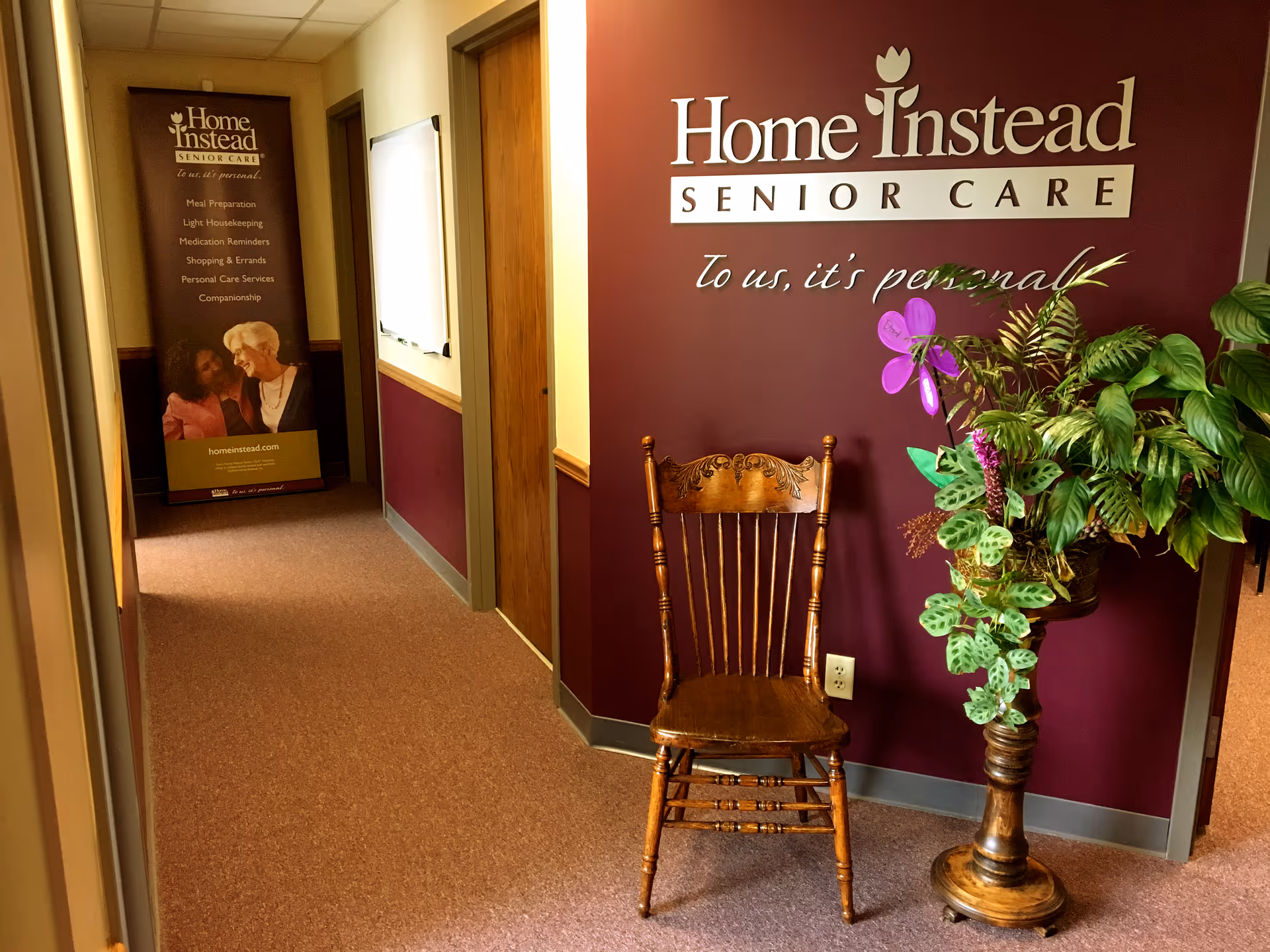 Maroon-walled hallway with a wooden chair and large plant beside a Home Instead sign.