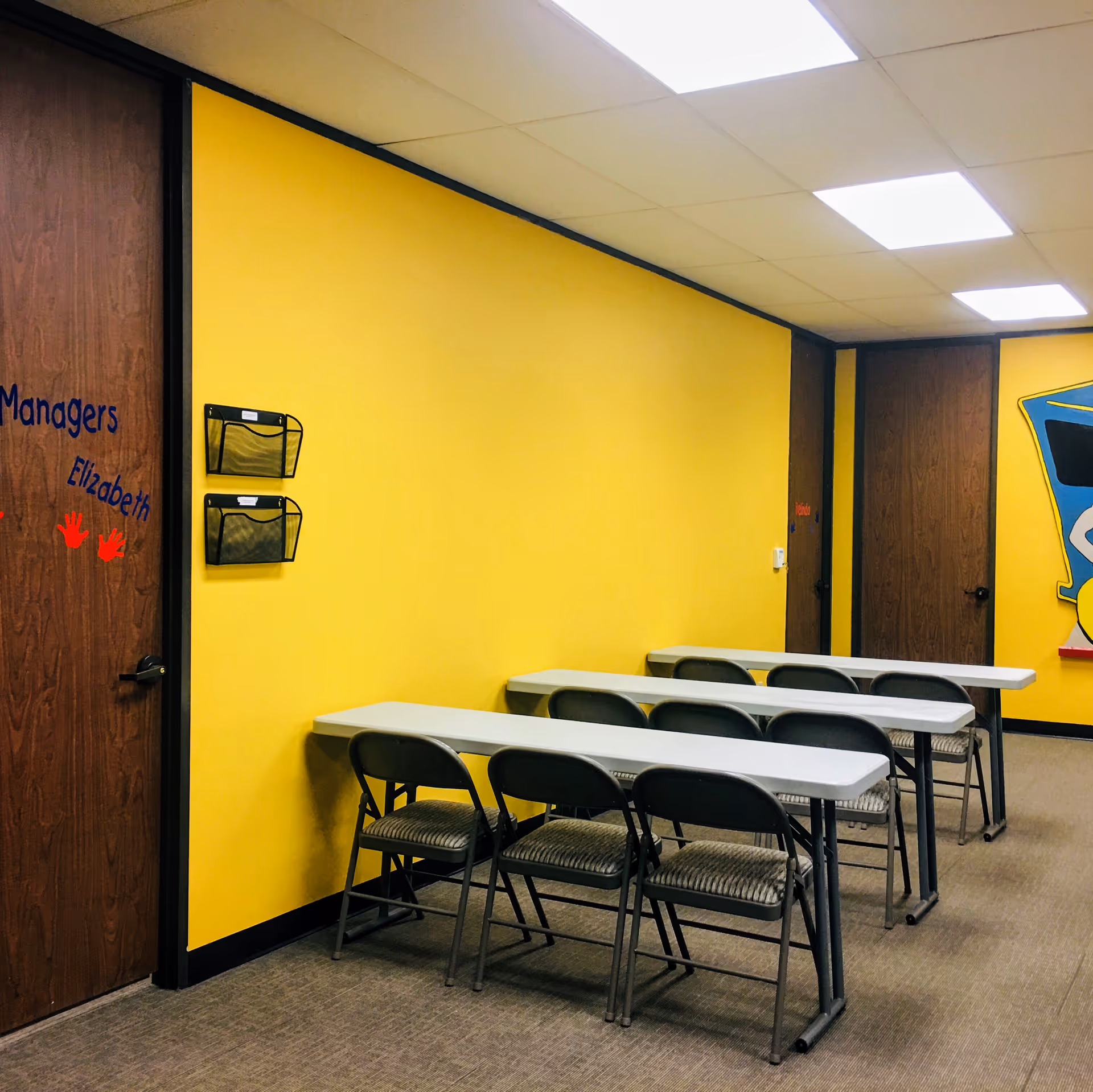 Yellow-walled classroom-style room with desks and folding chairs.