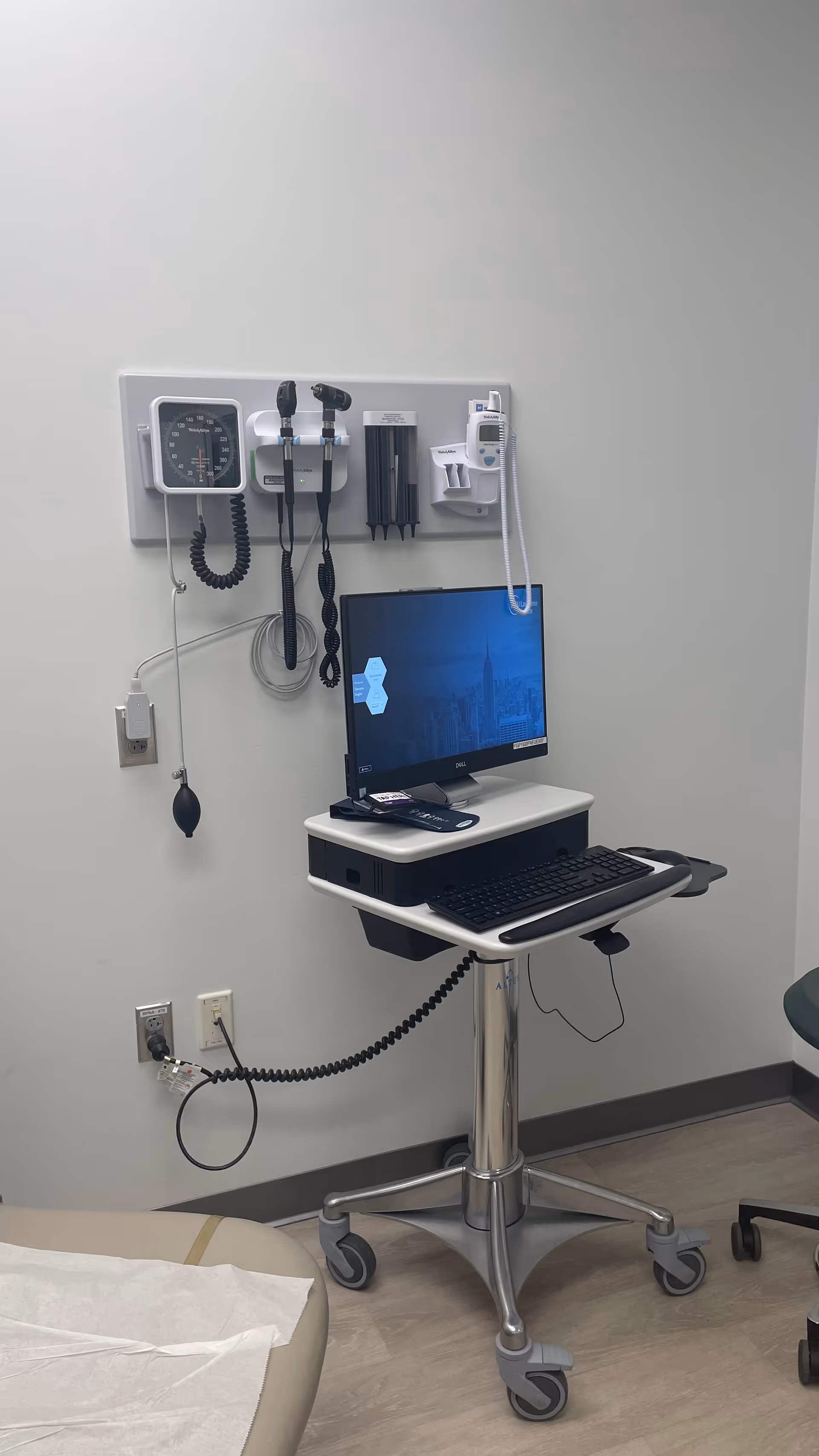 Medical exam station with wall-mounted monitor, blood pressure cuff, and mobile computer cart.