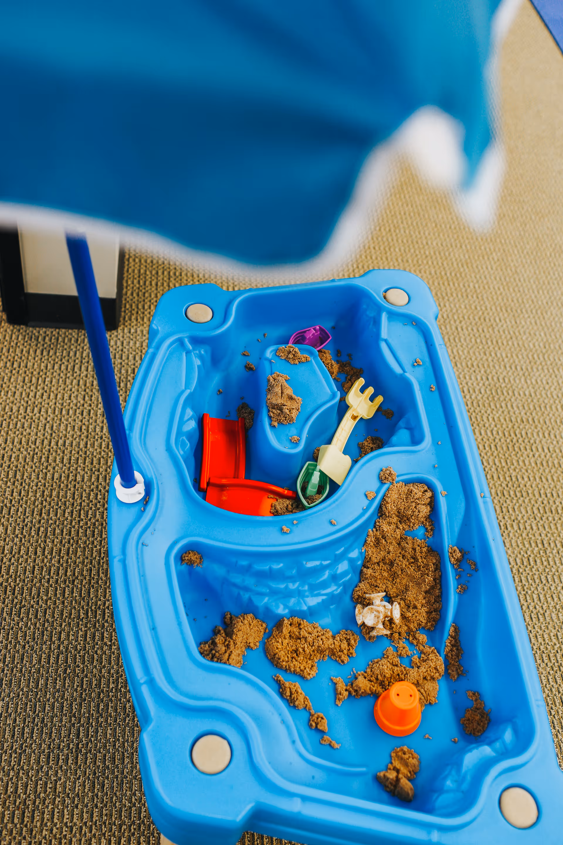 Blue plastic sand table with scattered sand and small toys on a carpeted floor.