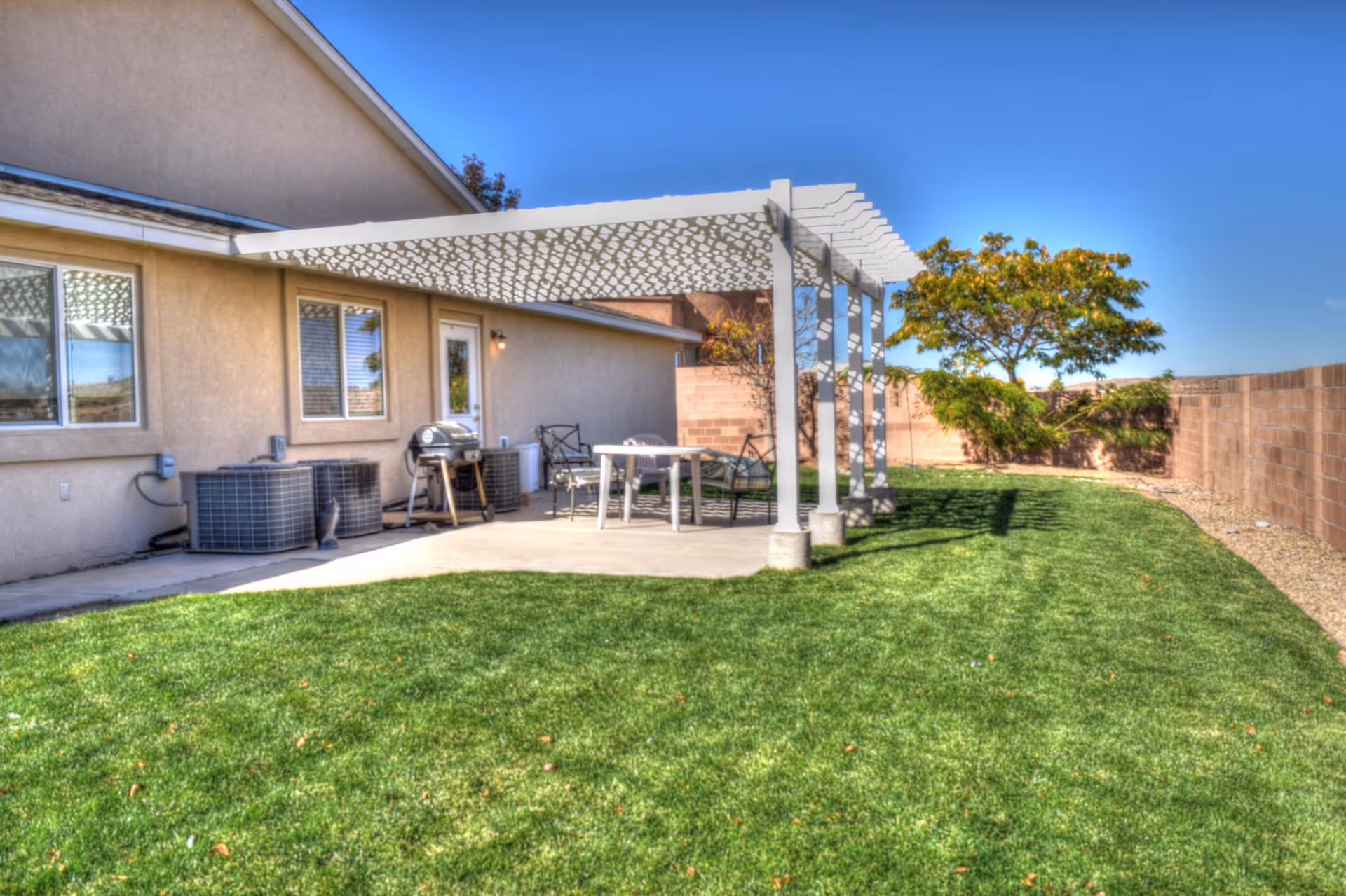 Outdoor patio with table, chairs, pergola, and green lawn beside a beige building.