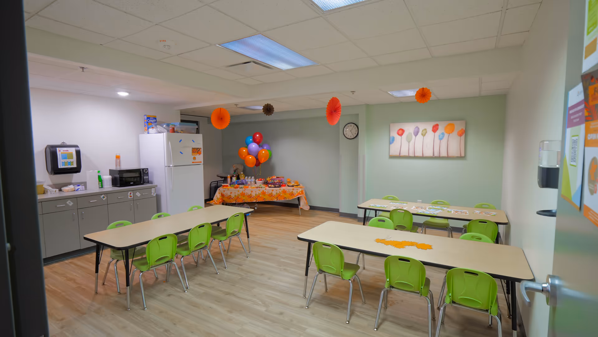Bright senior living dining area with green chairs around tables and a kitchenette corner.