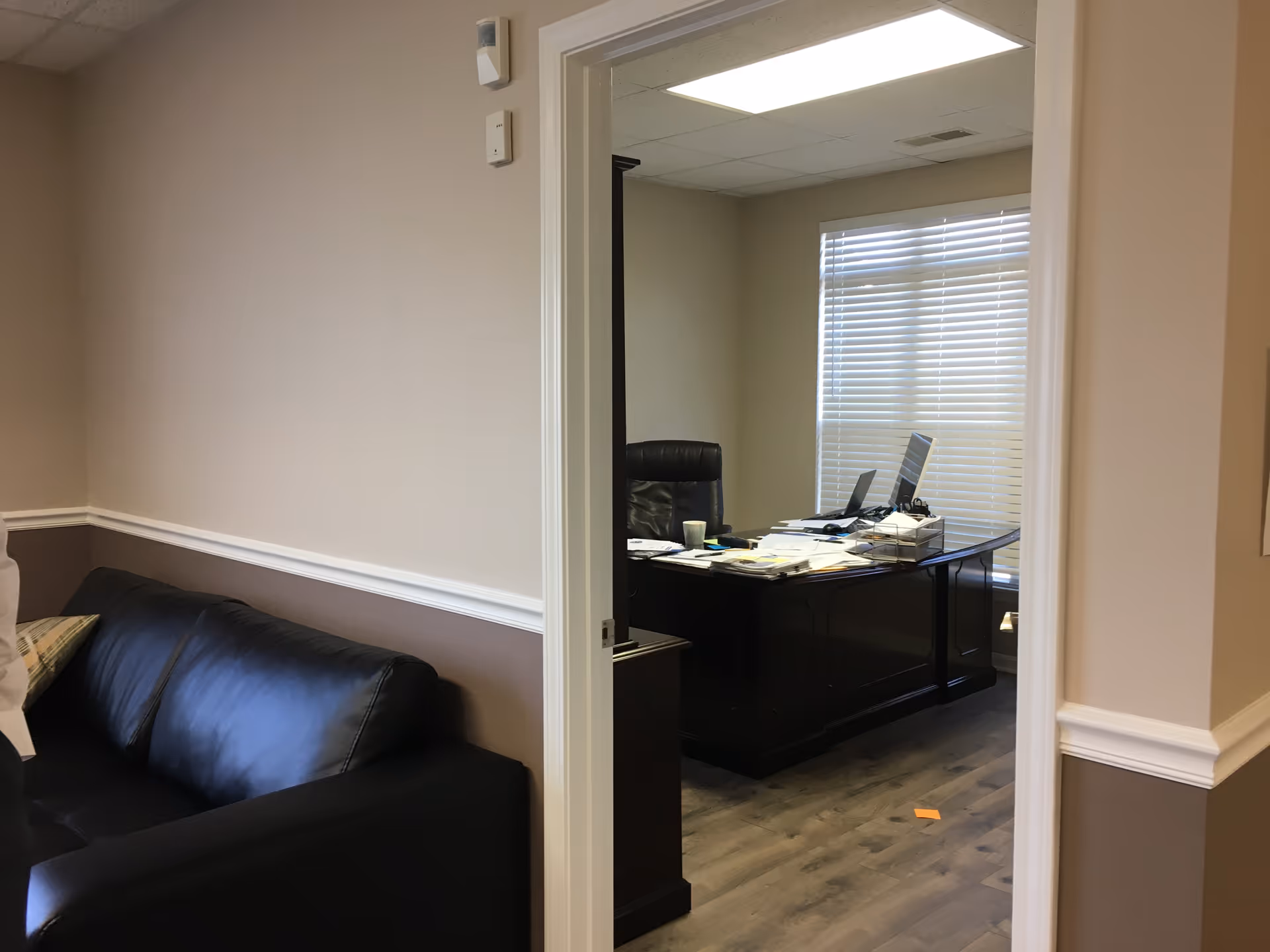Office workspace with desk, computer, papers, and a black leather couch in a beige hallway.