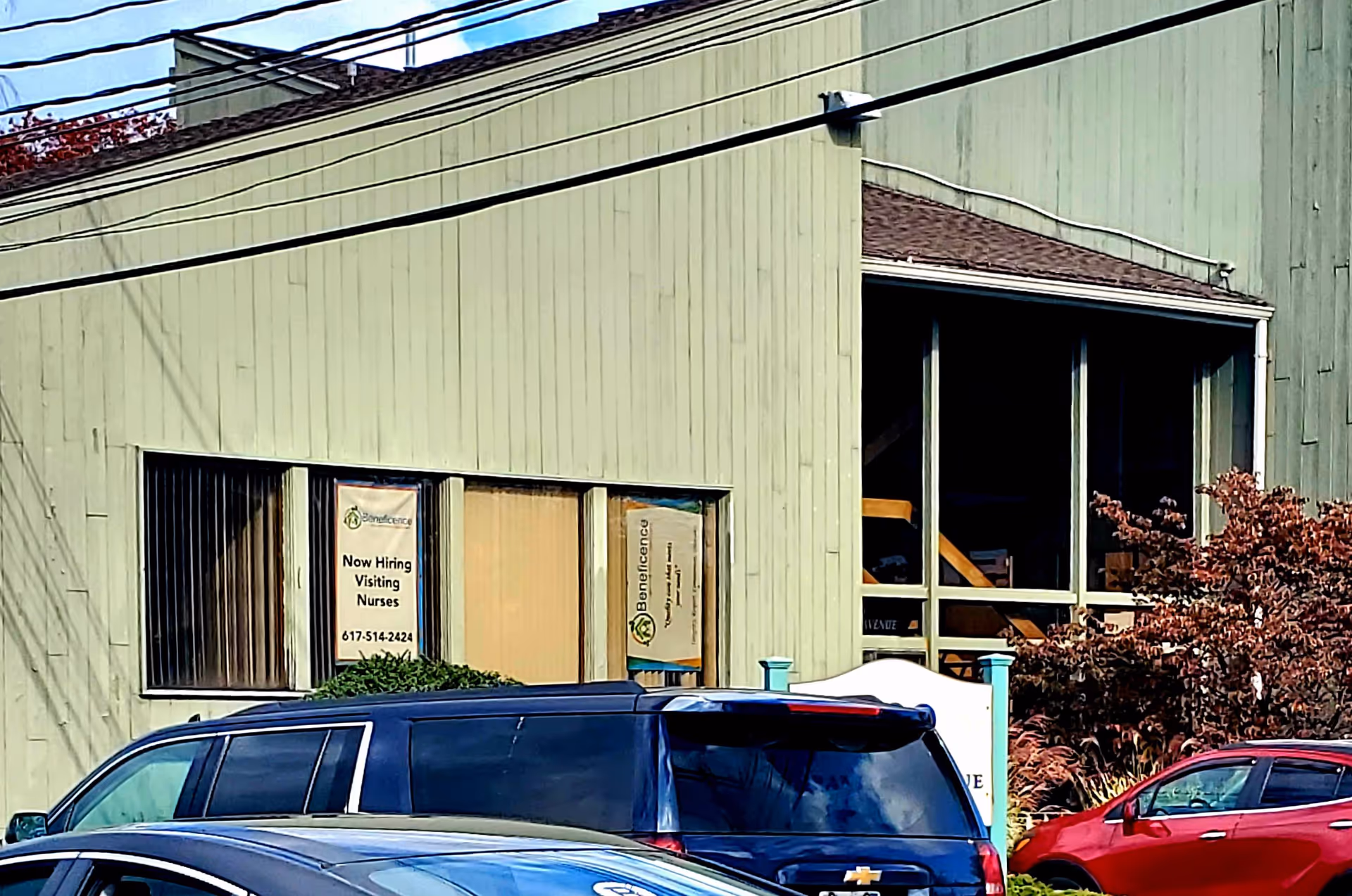 Exterior view of Beneficence Home Healthcare building with Now Hiring signs in windows and parked cars.