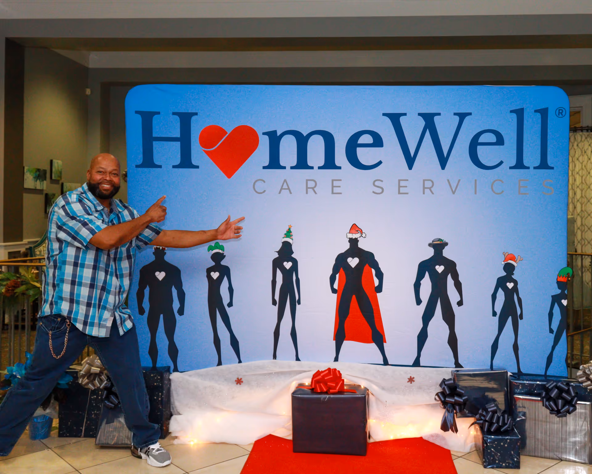Man in plaid shirt points at a large HomeWell Care Services banner inside a facility.
