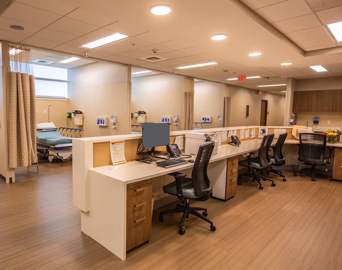 Nurse station with desks, computers, and curtained patient bays in a clinical interior.