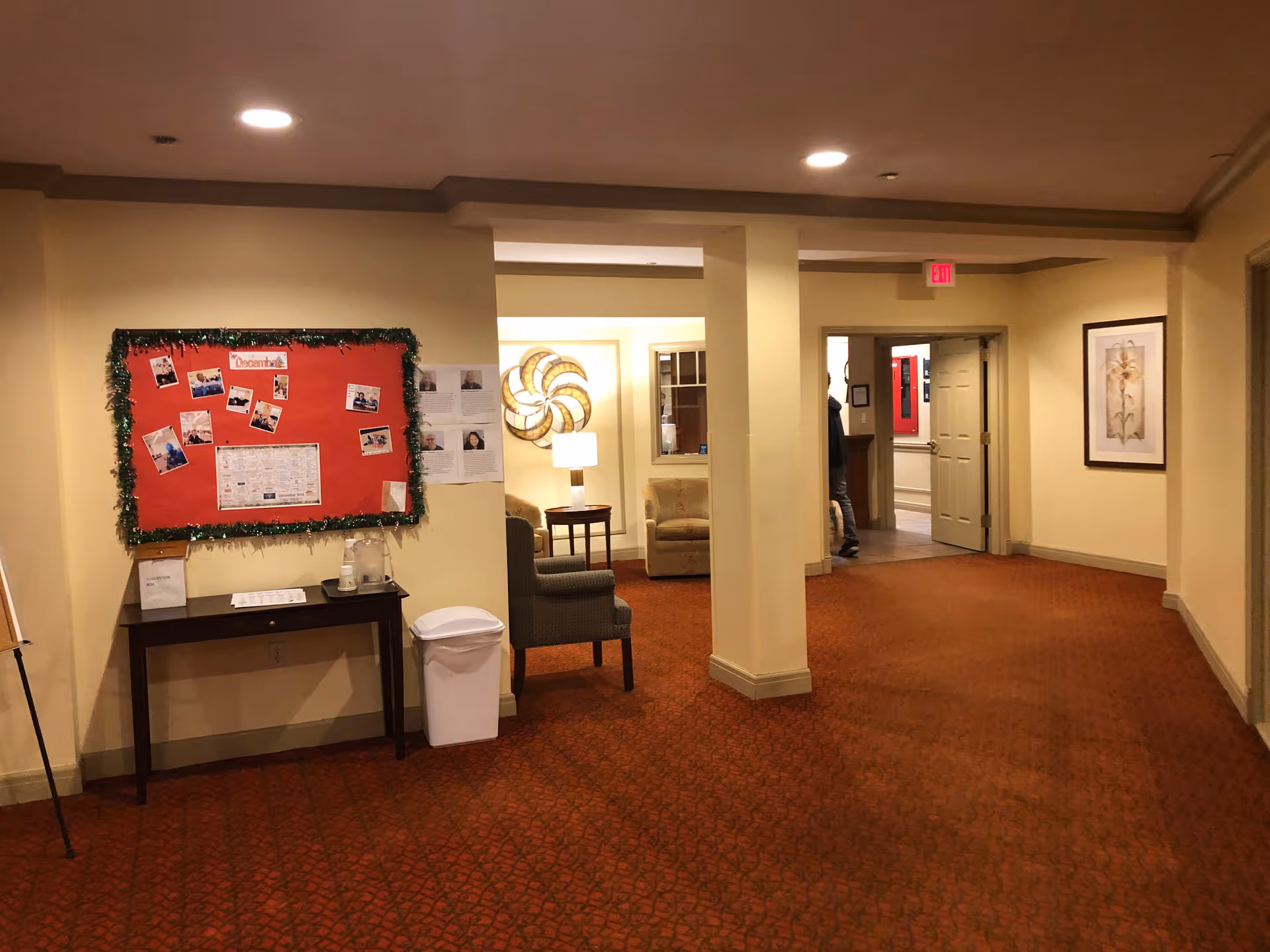 Interior hallway of a senior living facility with bulletin board and seating.