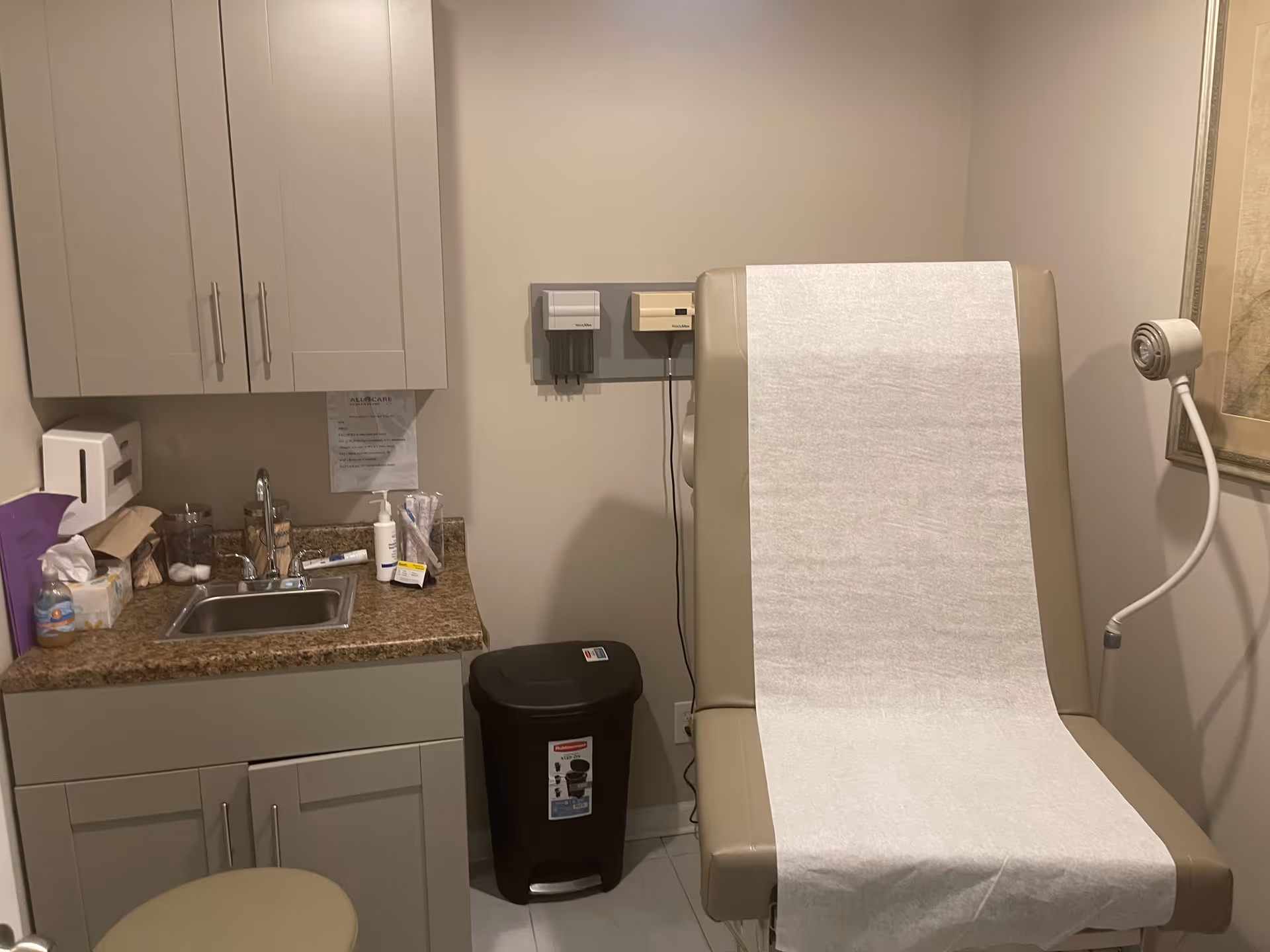 Exam chair covered with disposable paper beside a sink, cabinets, and medical supplies.