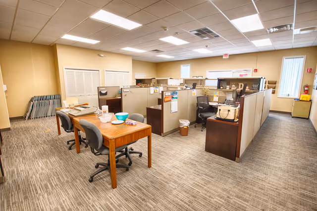 A spacious office area with cubicles, a wood table, and stacked chairs in a beige-walled facility.