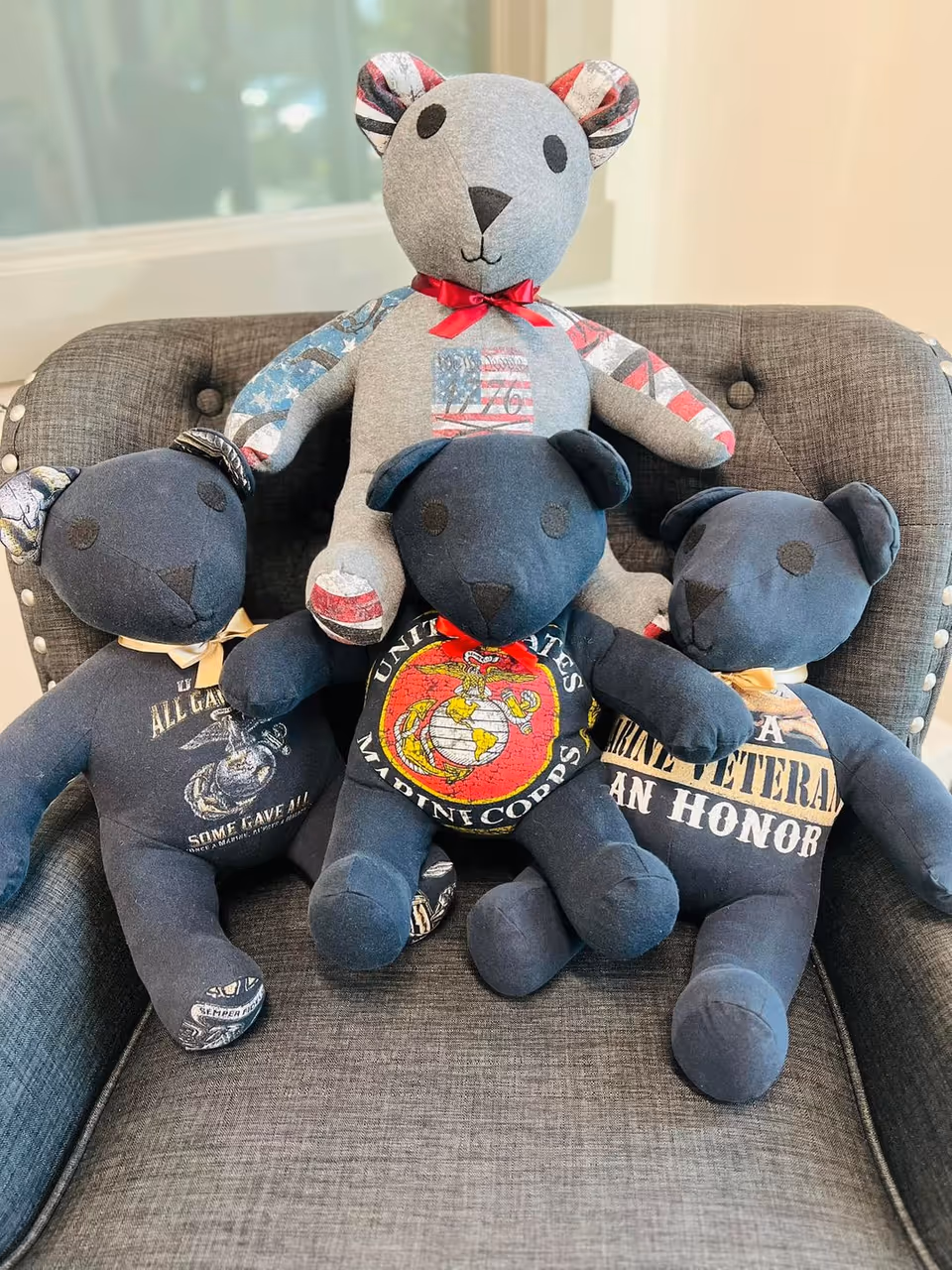 Three navy bears and one gray bear with patriotic patches sit on a gray couch indoors.