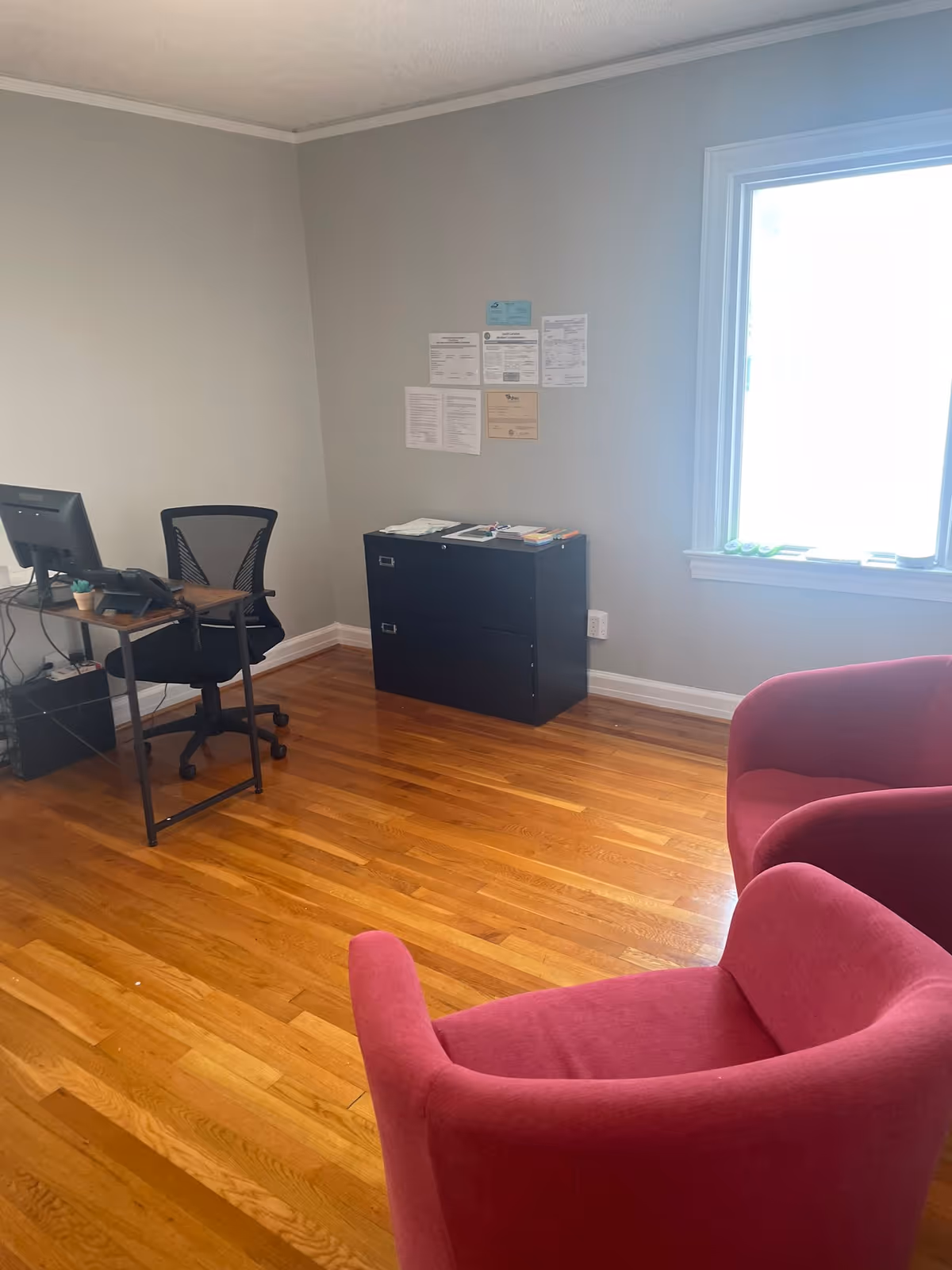 Office area with desk, computer, filing cabinet, and pink chairs by a window.