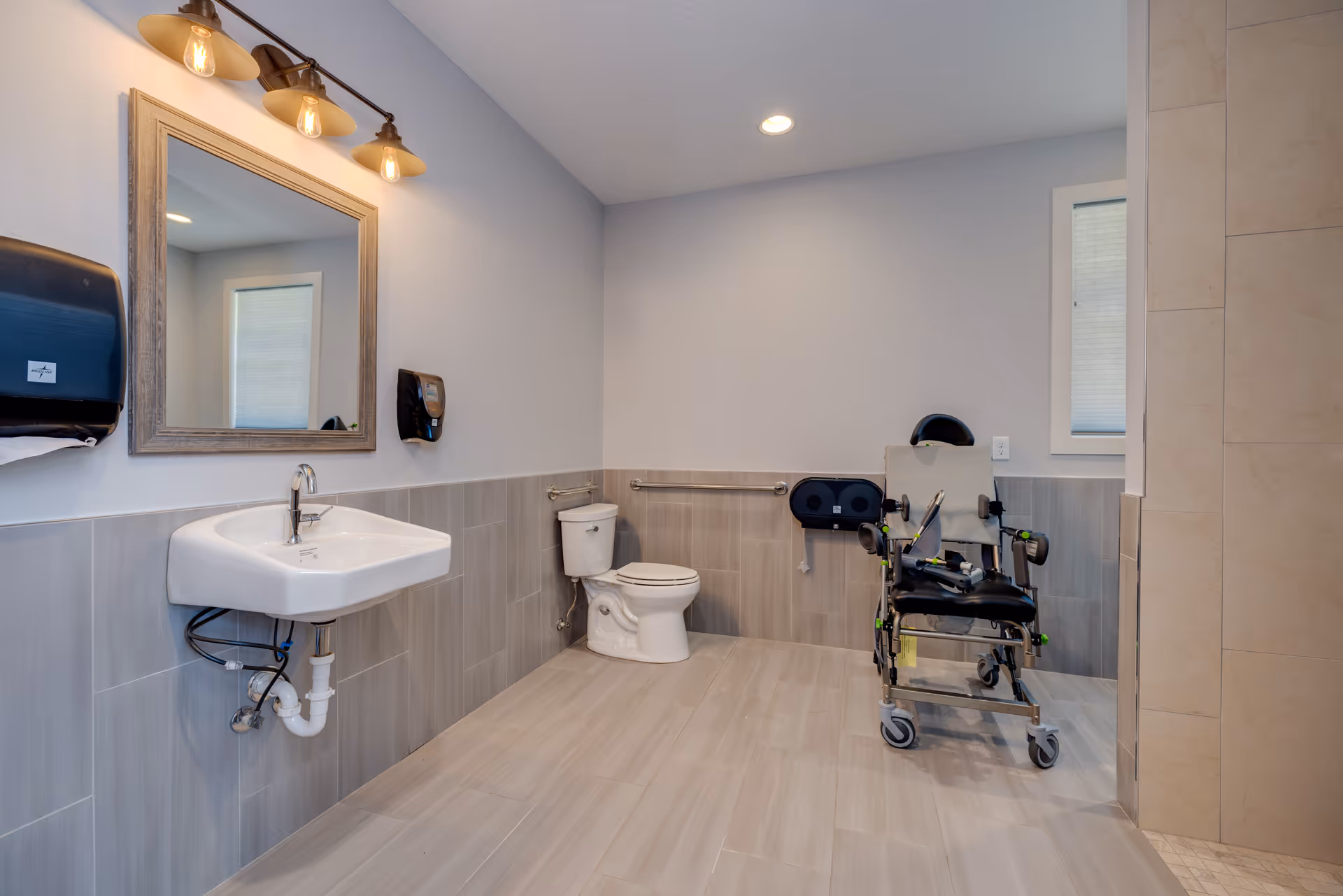 Clean bathroom with sink, mirror, toilet, and a wheelchair in a care facility.