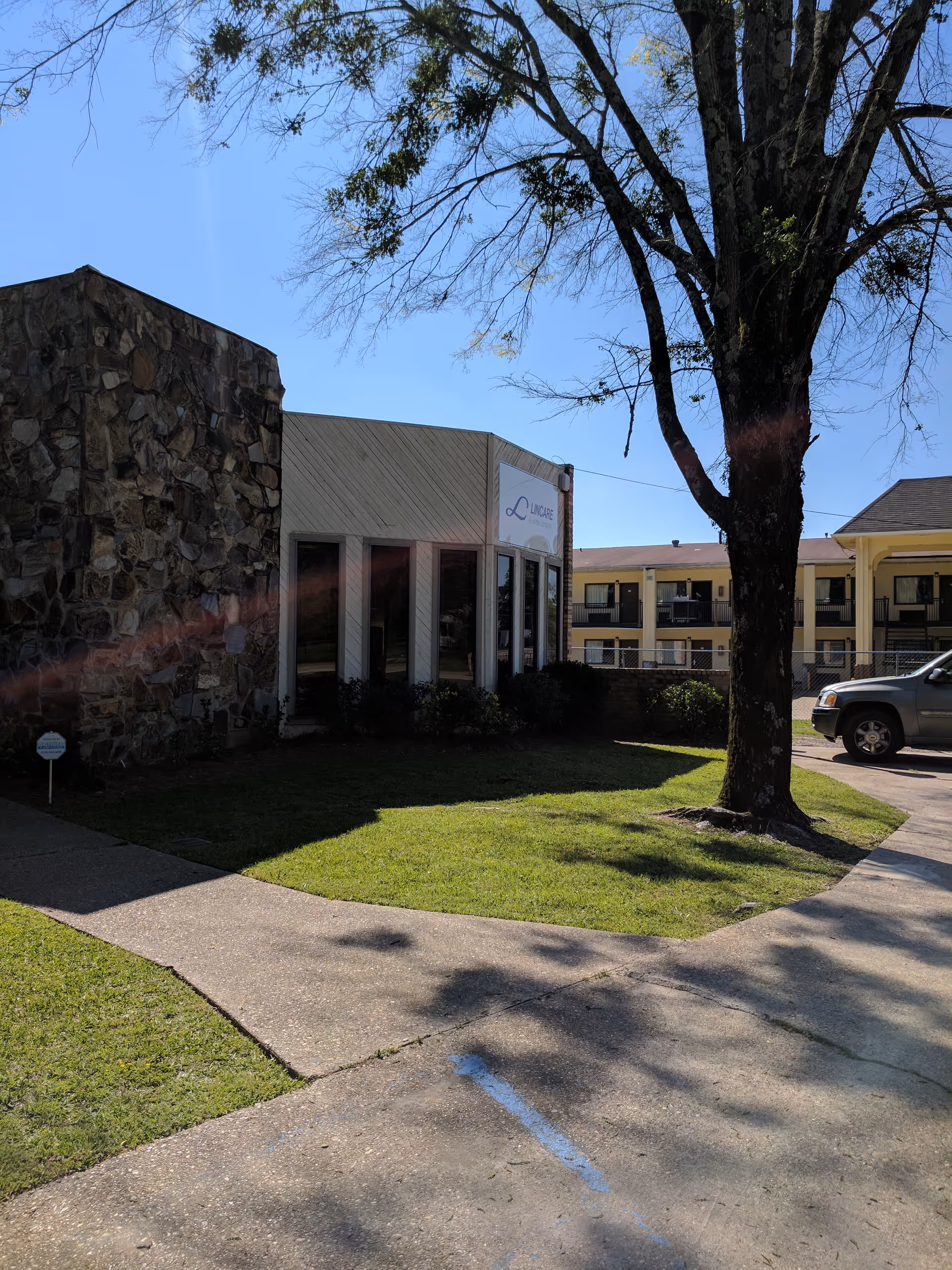 Exterior view of a LINCARE senior living building with a tree, lawn, and parked car.