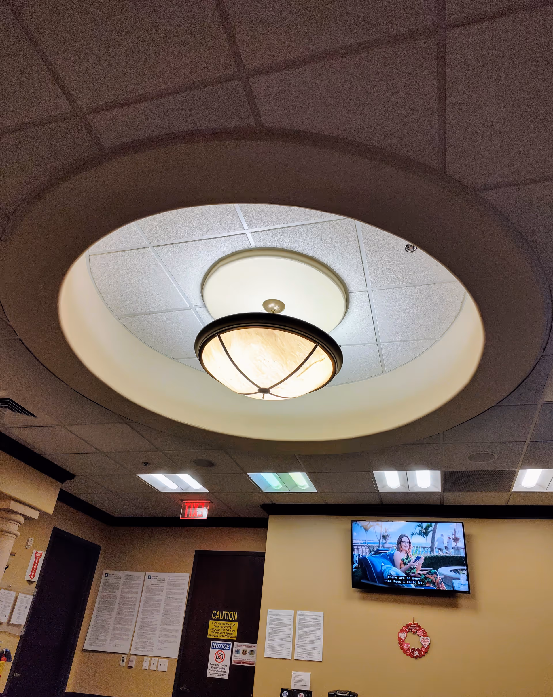 Circular ceiling light fixture looms over a hallway with a wall TV and posted notices.