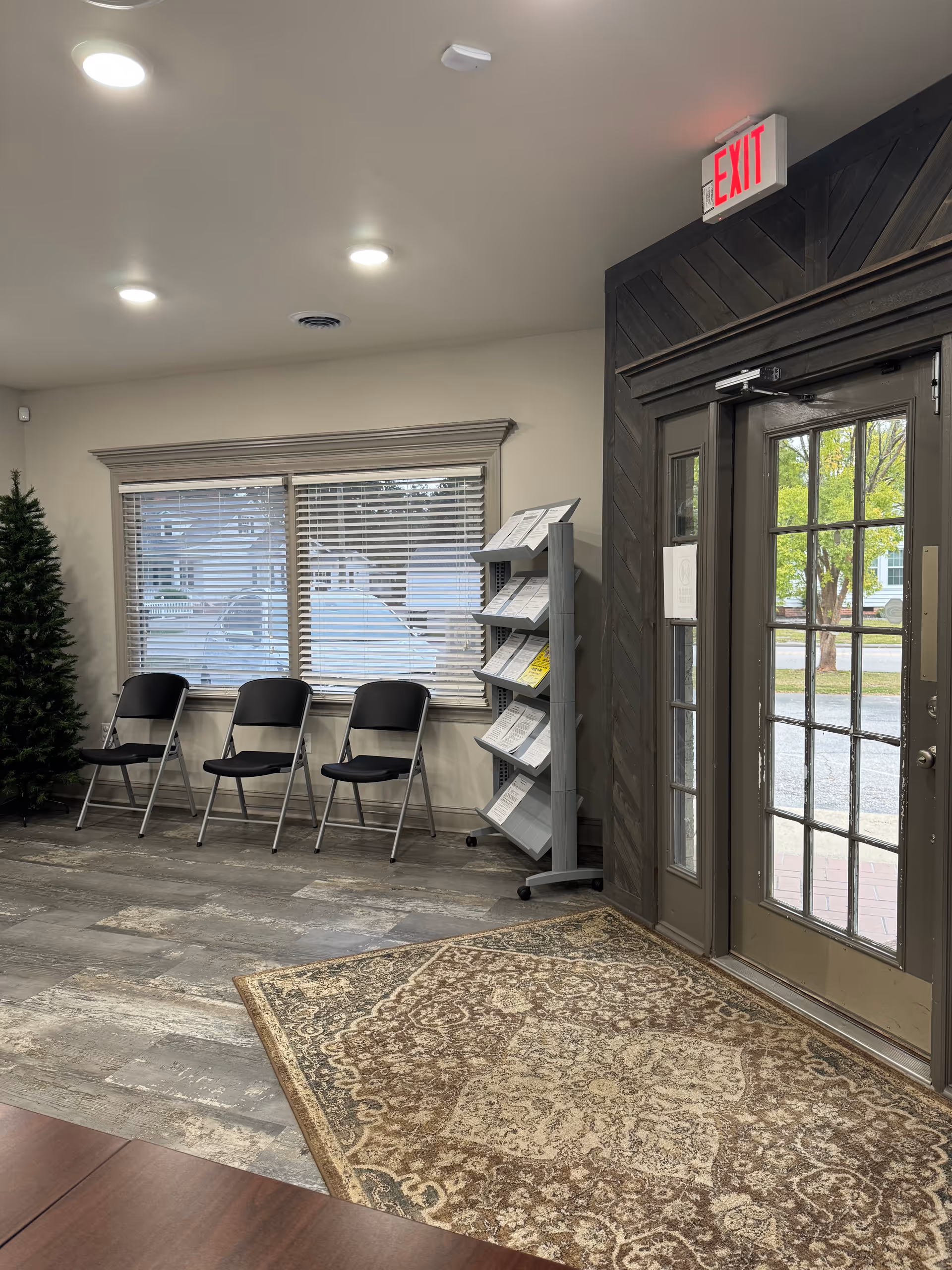 Interior lobby with four black chairs, brochure rack, rug, and glass exit door.