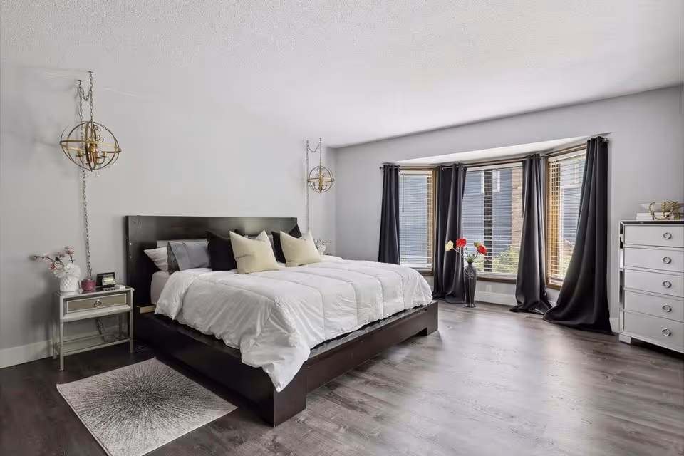 A modern bedroom with a large bed, nightstands, dresser, and bay windows with dark curtains.