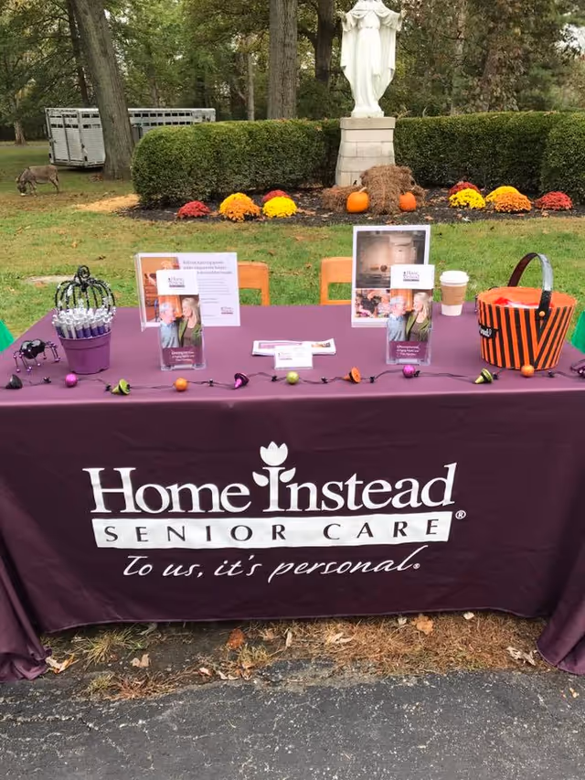 Outdoor event setup with a Home Instead bannered table, pamphlets, pumpkins, and fall decorations.