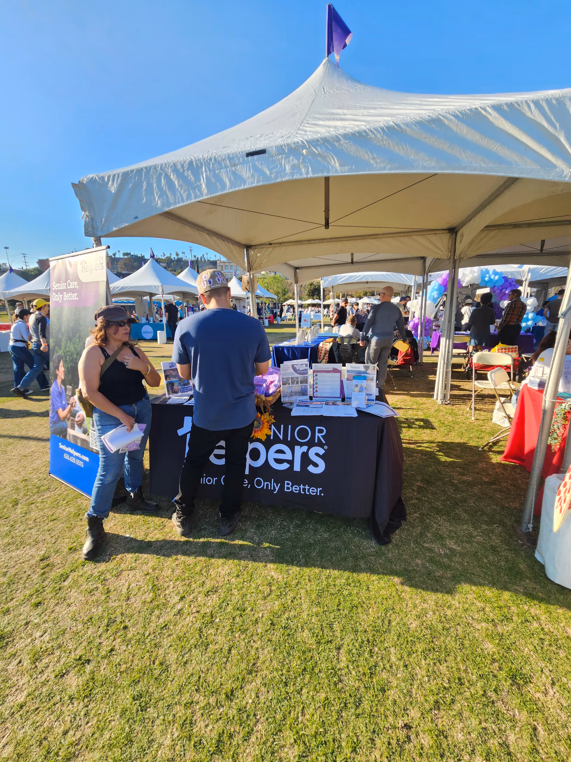 Senior Helpers booth at an outdoor event with tents, tables, and attendees
