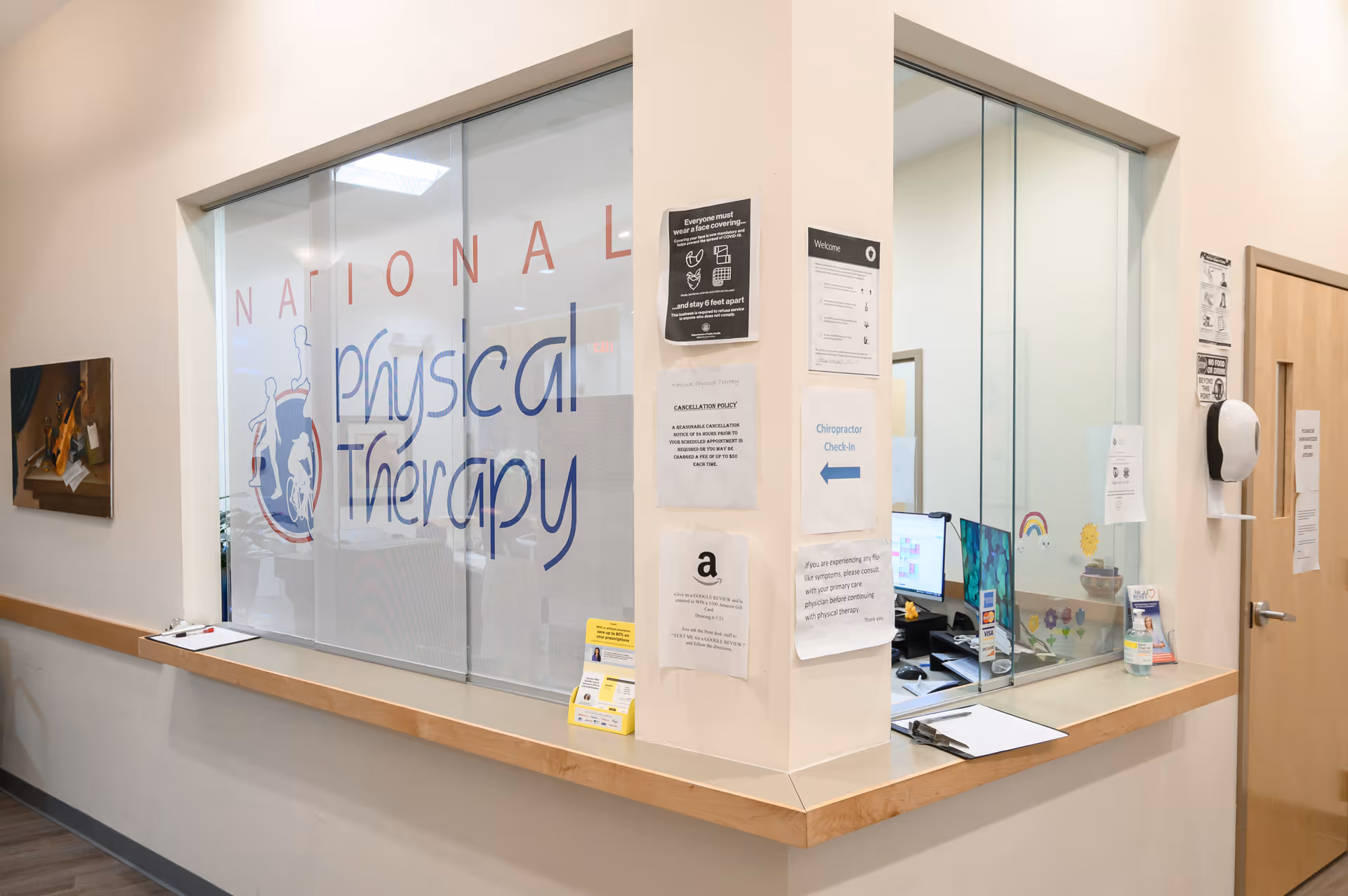 Reception desk inside a National Physical Therapy clinic, with glass panels and posted notices.