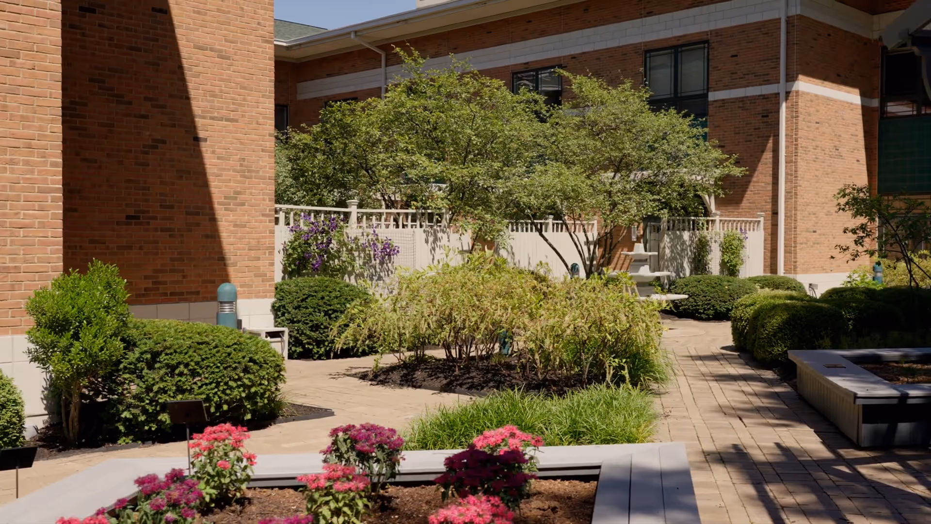 Outdoor courtyard at a brick senior facility with trees, shrubs, and colorful flower planters.