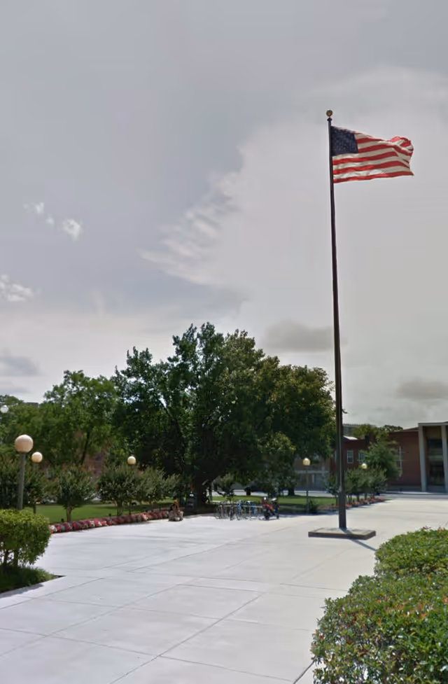 A wide outdoor courtyard with a tall flag, trees, and a modern building in background.