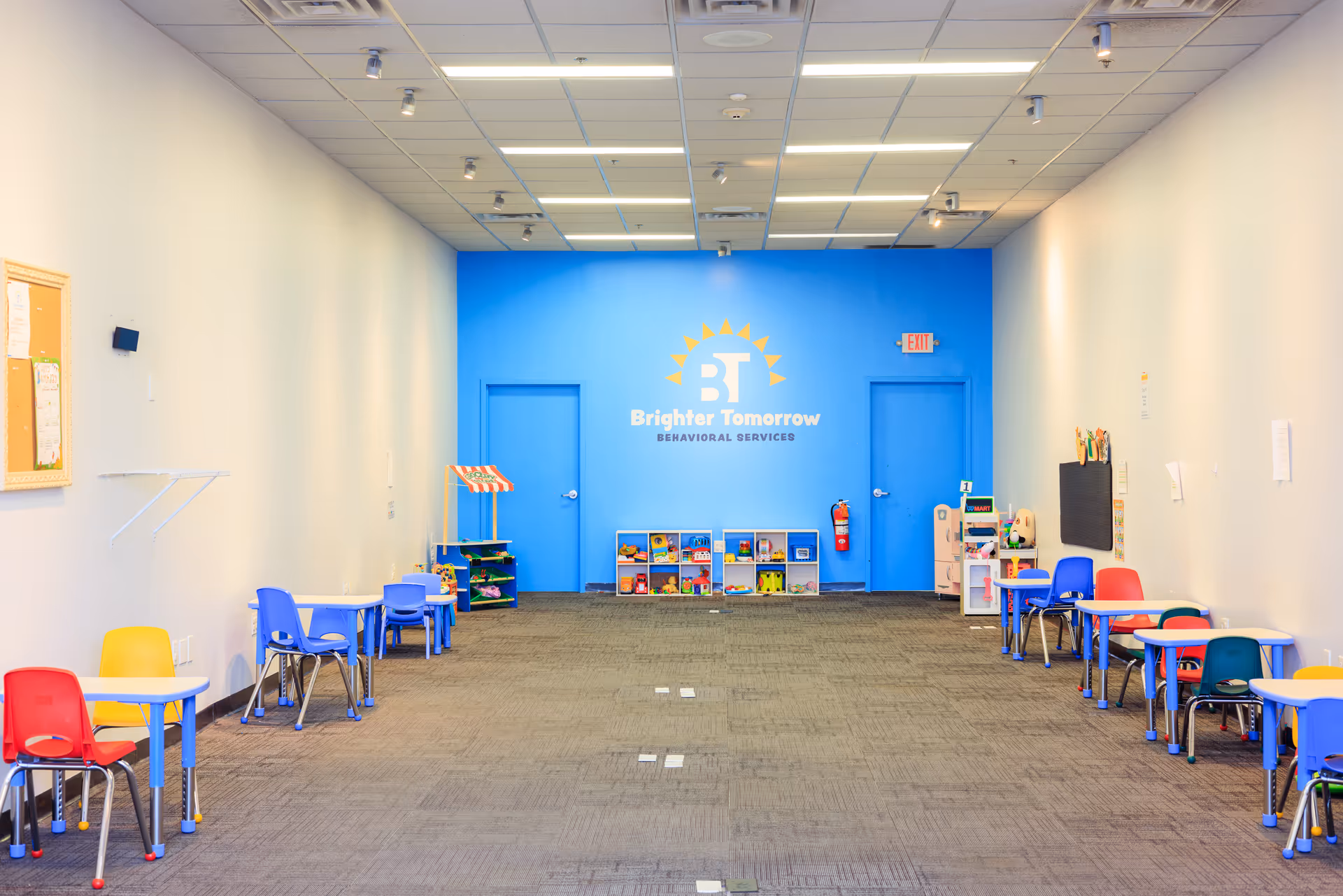 Bright activity room with blue branded wall, colorful chairs, tables, and toy storage along sides.