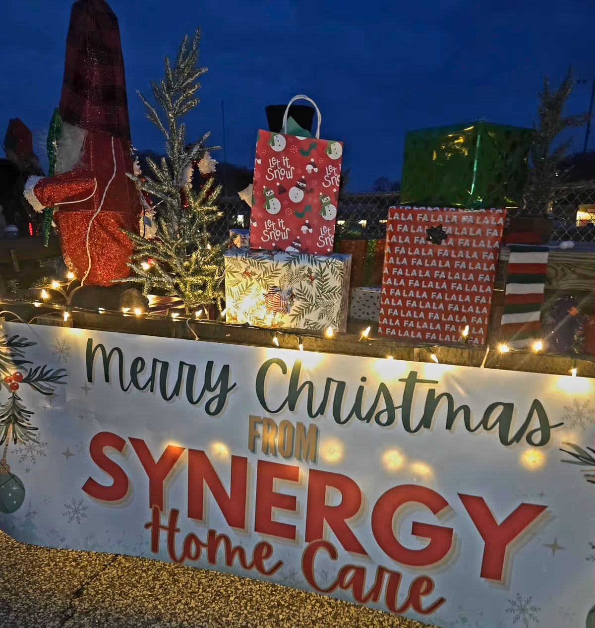 Nighttime exterior holiday display with wrapped gifts and a SYNERGY Home Care banner.