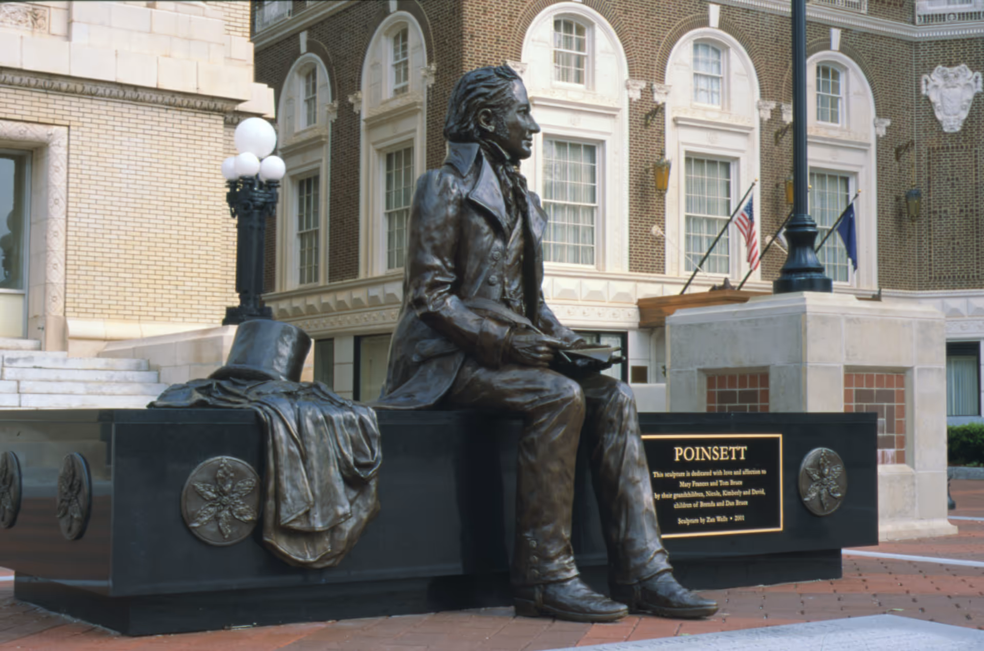 Bronze statue of a seated man in a city plaza with building and flags.