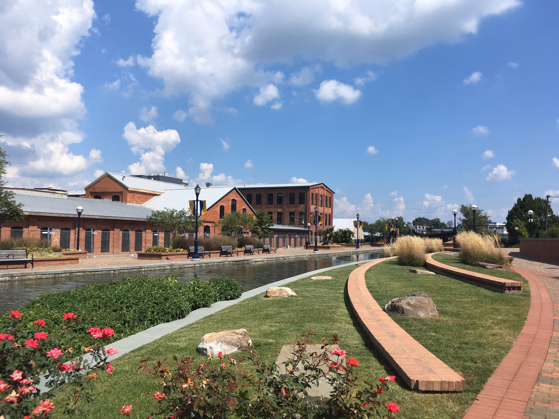 Sunny outdoor courtyard with brick buildings, benches, a water feature, curved brick paths, and landscaped greenery.