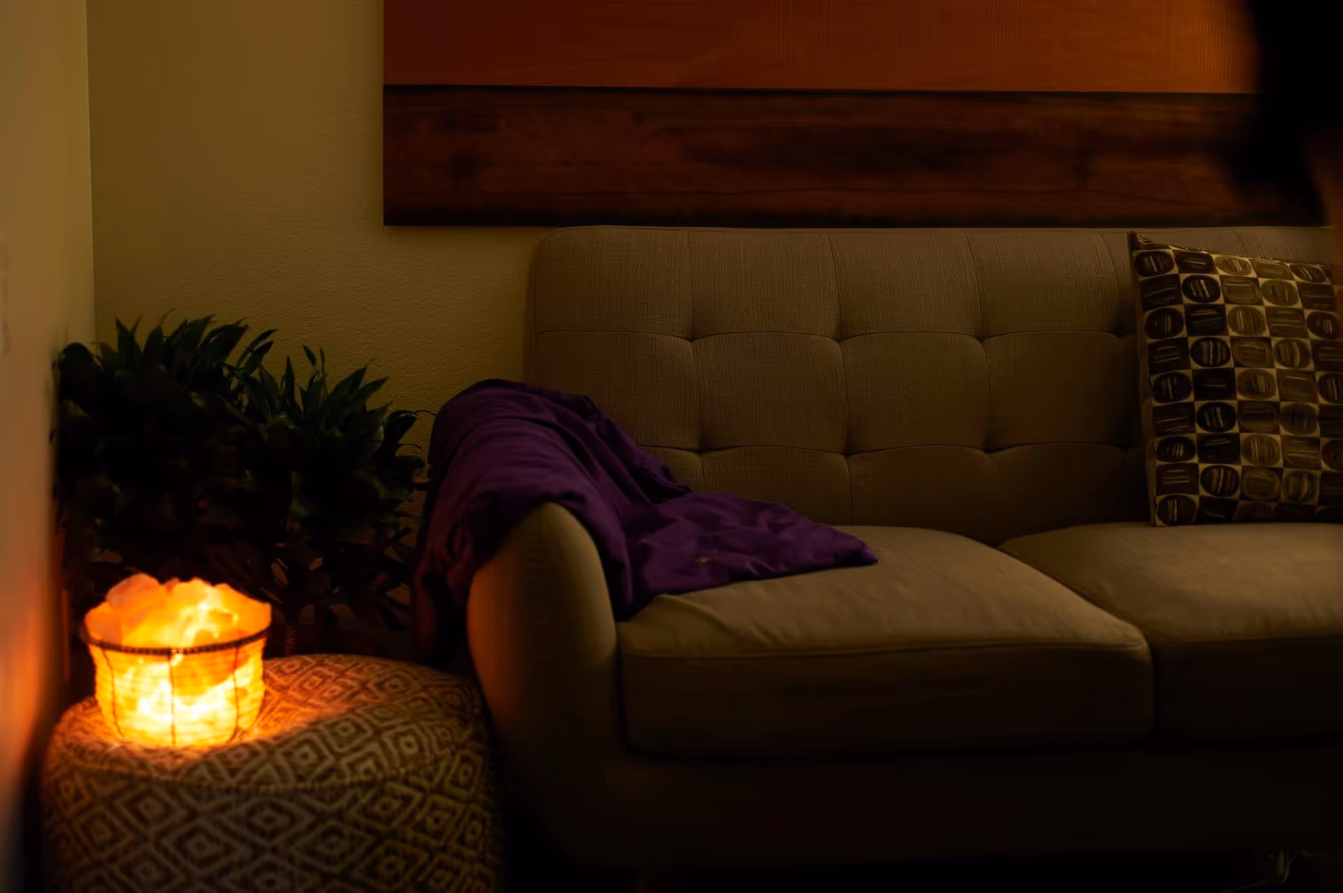 Dimly lit living room with beige sofa, purple blanket, pillow, salt lamp, and plants.