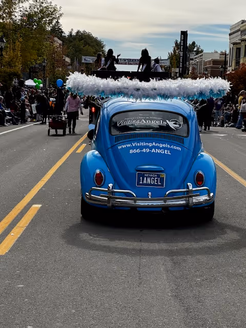 Blue vintage car with Visiting Angels signage drives through a crowded parade.