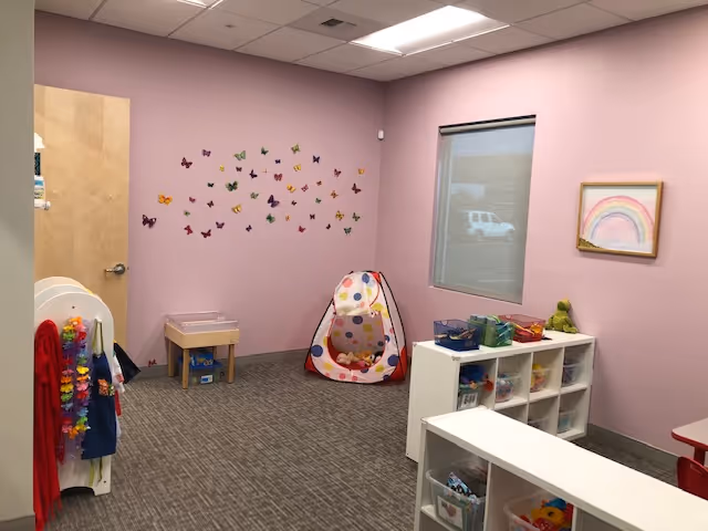 Interior pink therapy playroom with butterfly decals, toys, cubbies, and a tent.