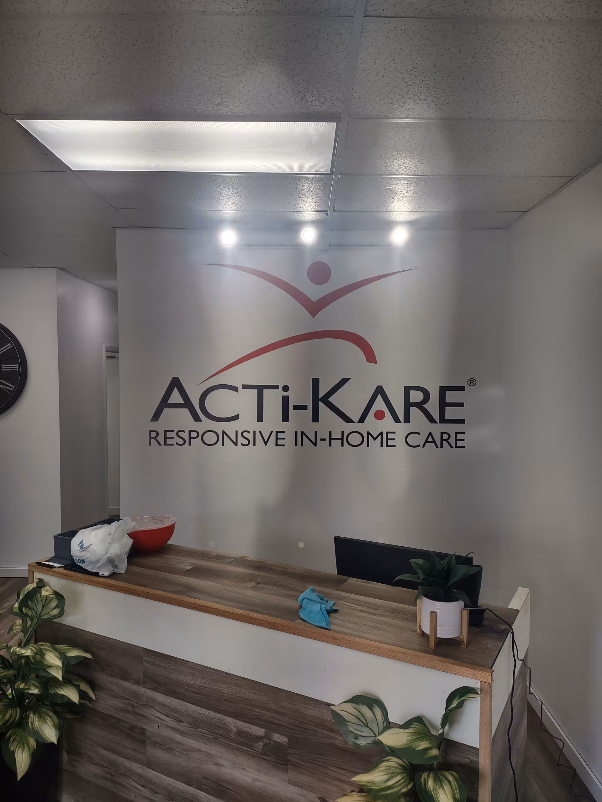 Reception desk with Acti-Kare logo on wall, plants, and office items.