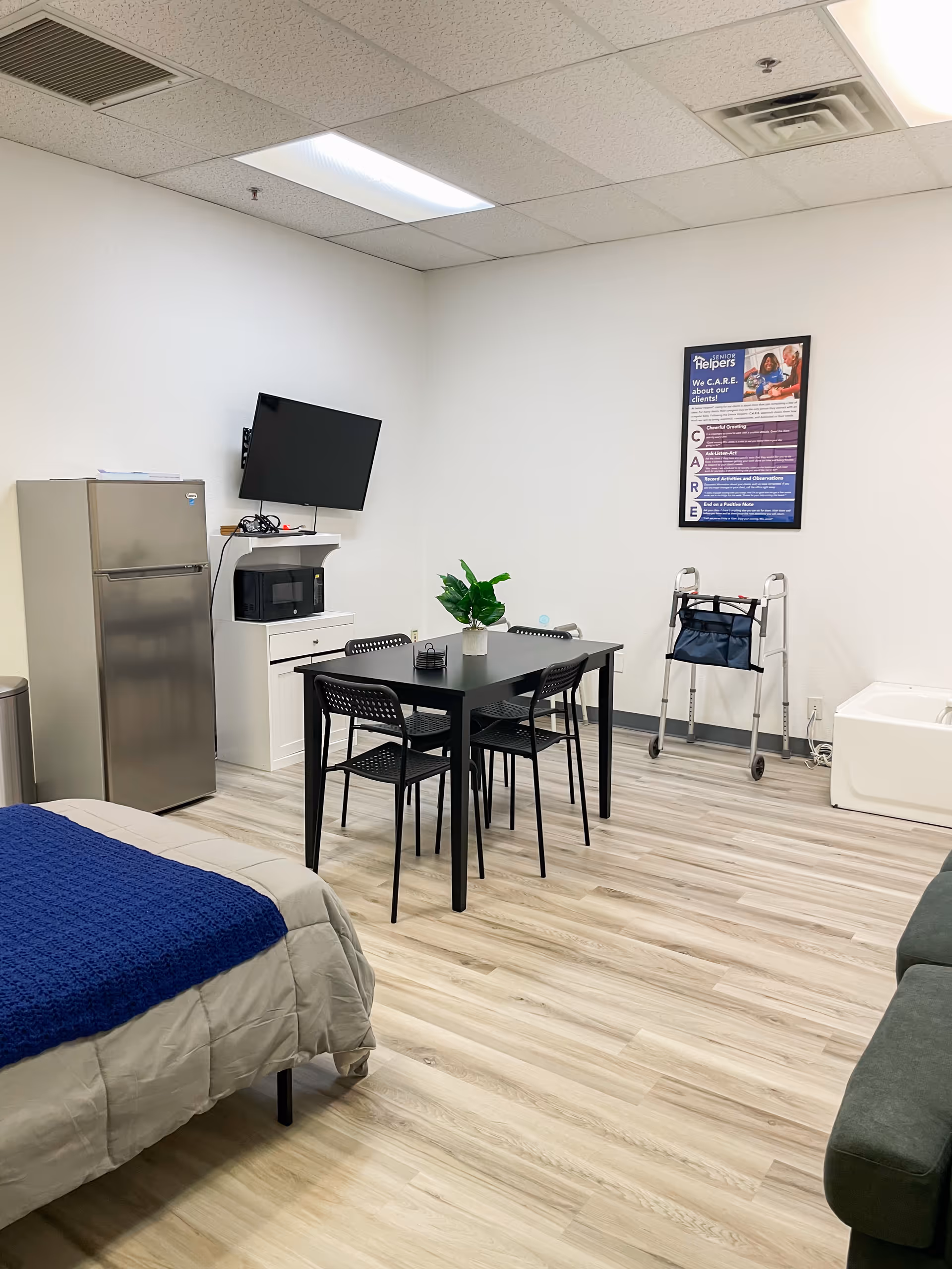 A small, bright facility room with a bed, dining table, fridge, TV, and walker.