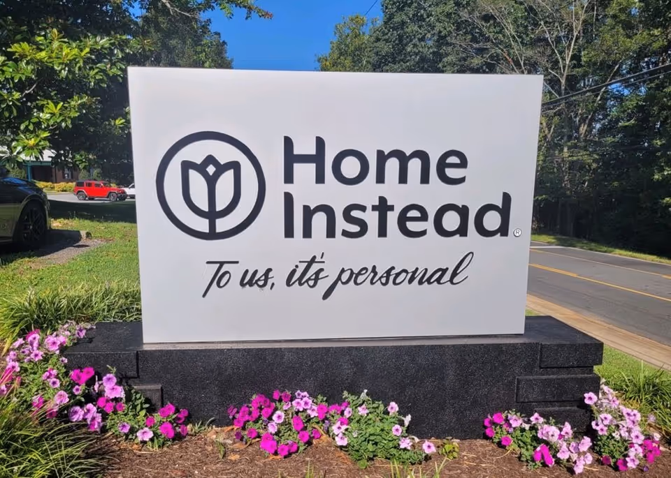 A large Home Instead sign on a stone base with flowers beside a road.