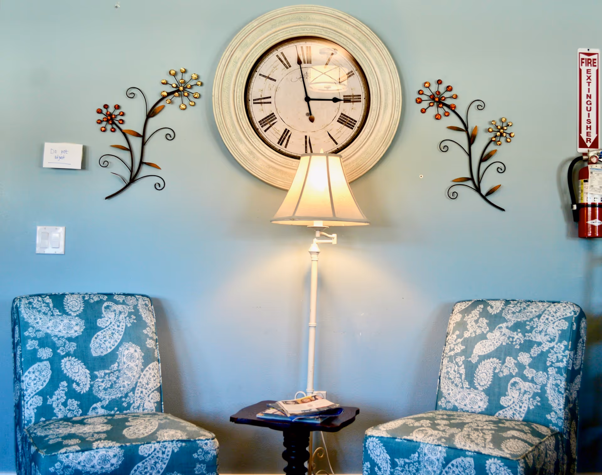 Two blue patterned chairs face a lamp and large clock on a blue wall.