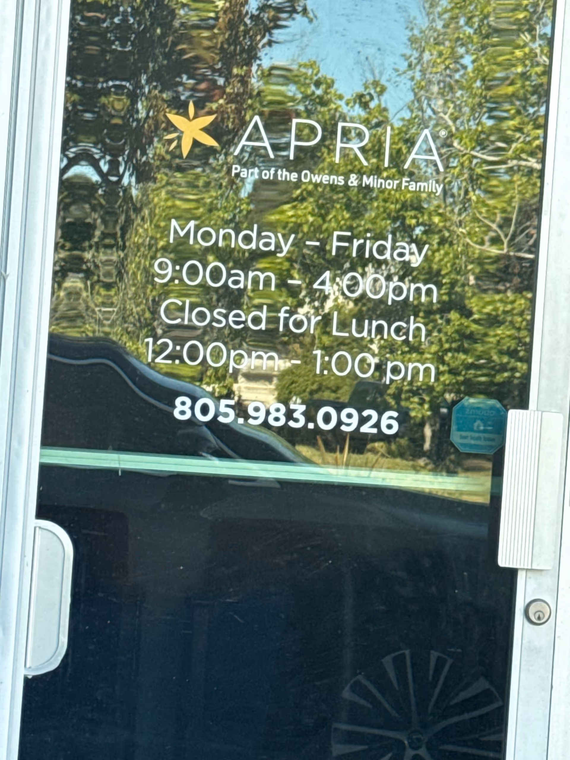 Glass door with Apria Healthcare logo and hours, reflecting trees outside.