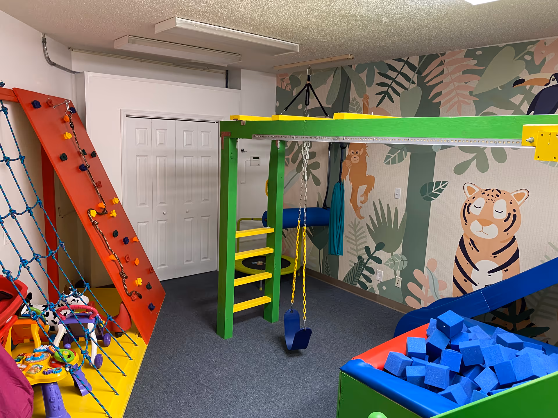 Colorful indoor play area in a senior facility with jungle mural, climbing wall, and foam blocks.