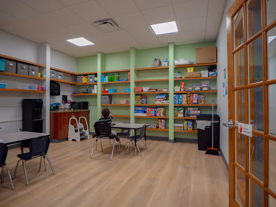 Interior playroom with green shelves, toys, and small tables; a child sits at a table.