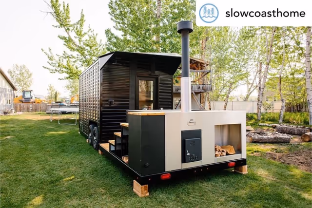 A black wooden sauna on a trailer with a stove sits in a grassy backyard.