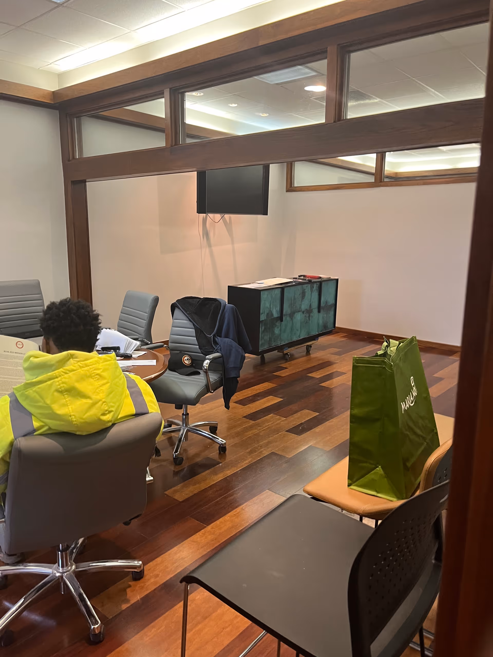A person in a neon jacket sits at a round table in a modern office-like room.