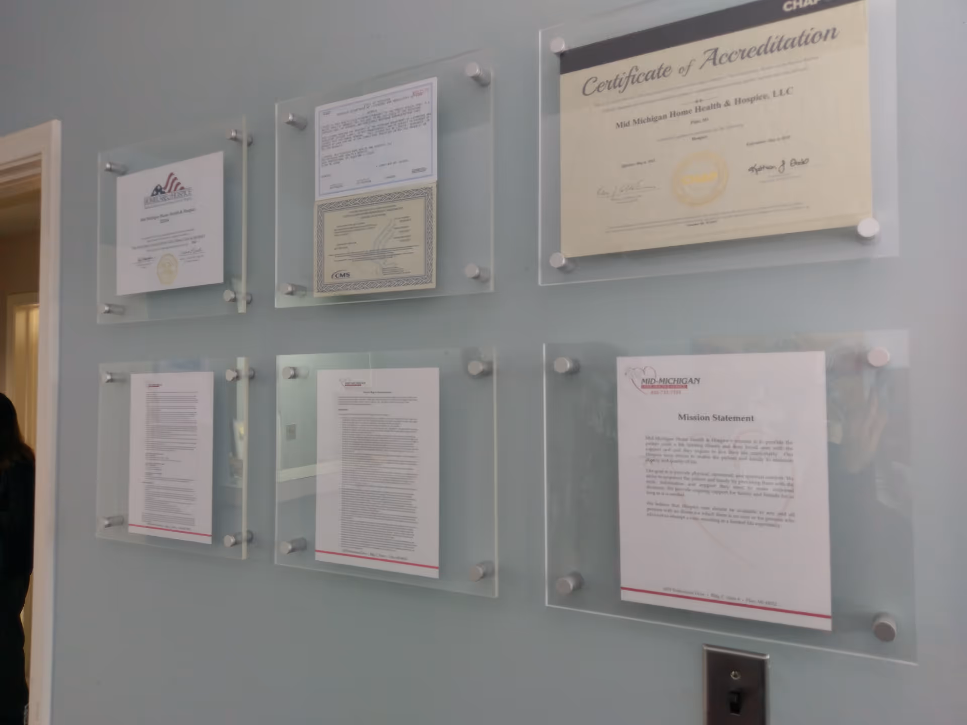 Glass plaques with framed certificates mounted on a light wall in a facility hallway.