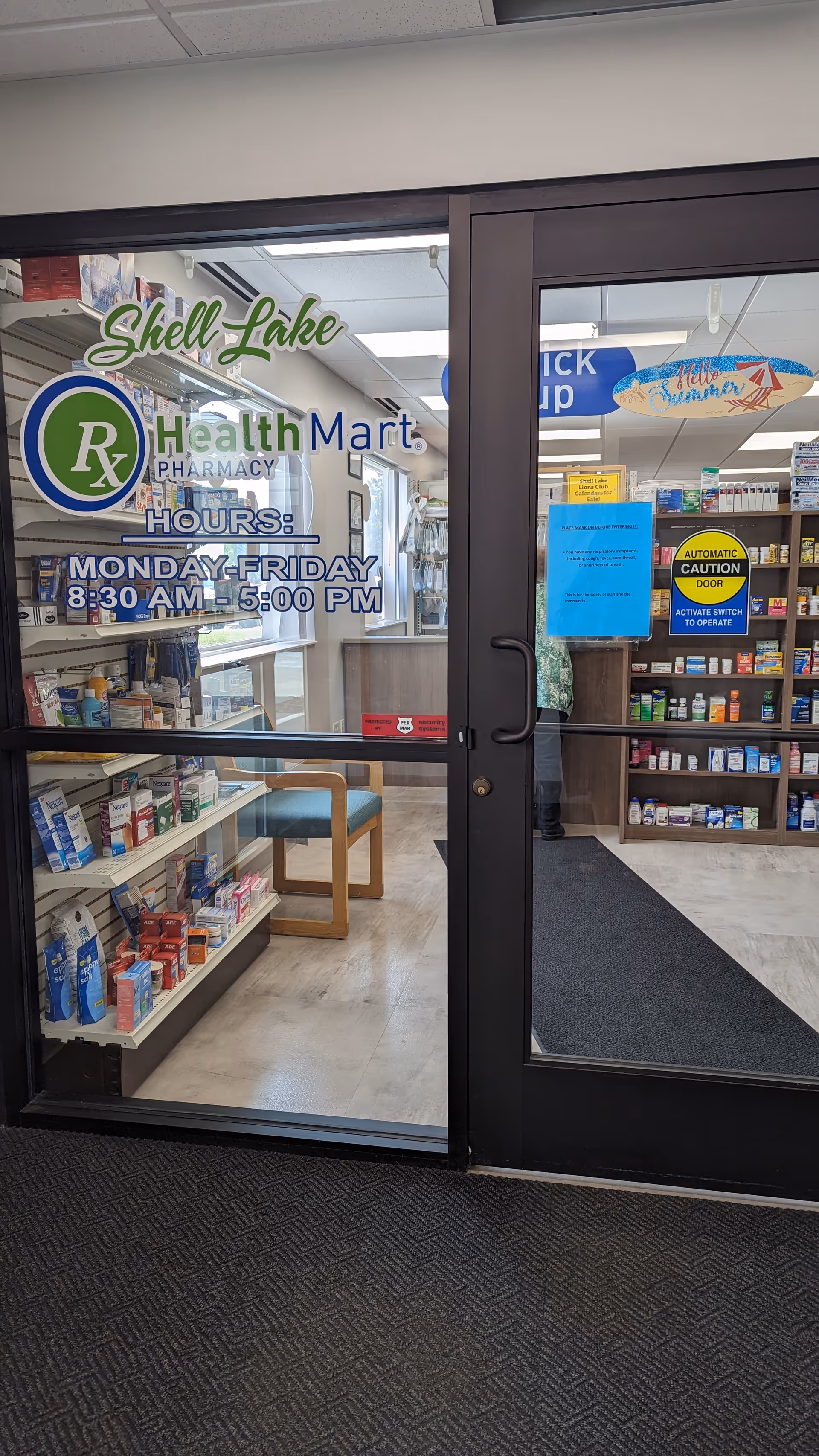 Glass entrance to a pharmacy with Shell Lake Health Mart hours and shelves inside.