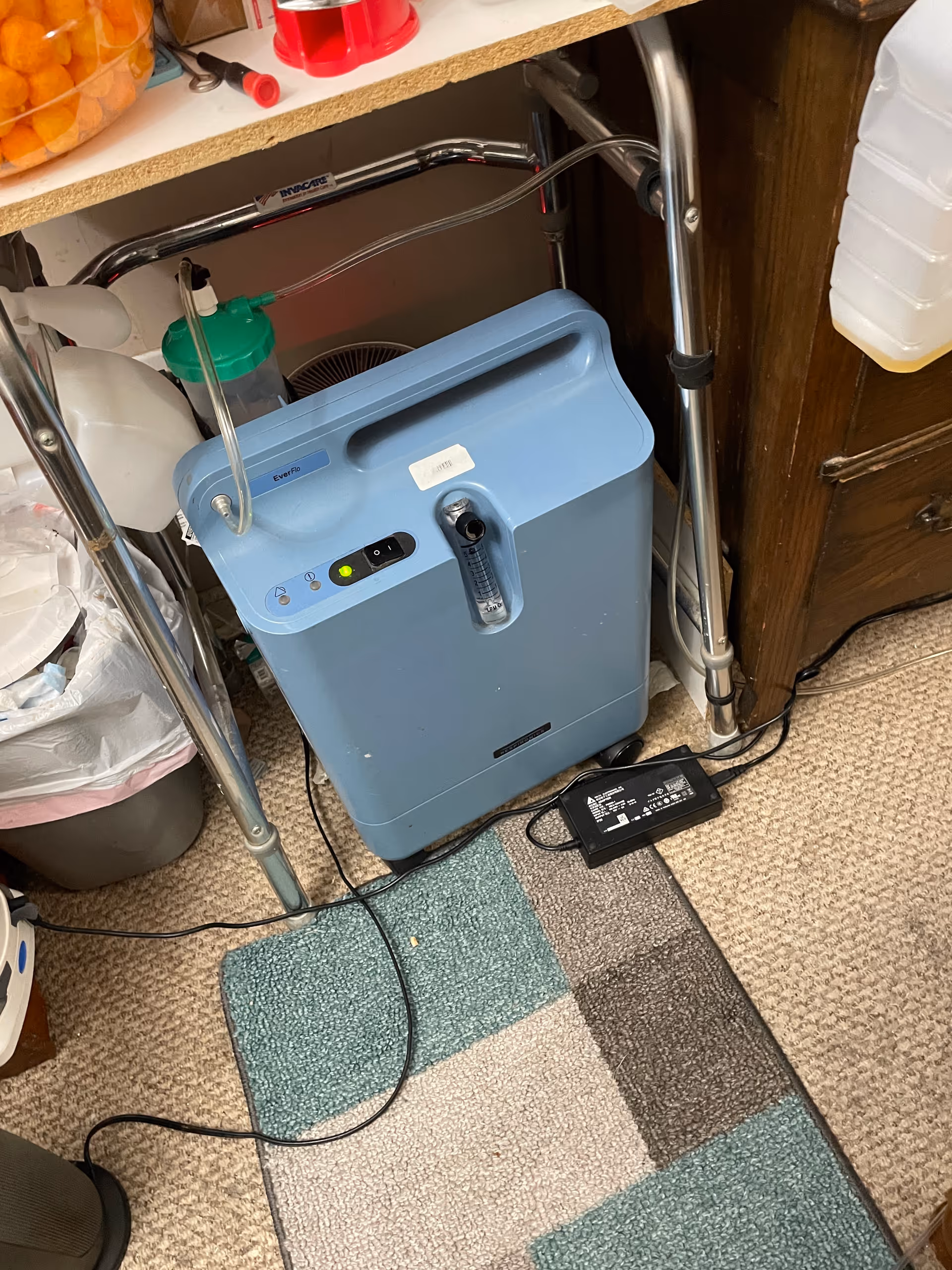 Blue oxygen concentrator with tubing under a shelf in a cluttered indoor utility space.