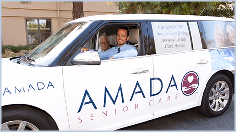 A white Amada Senior Care van with two smiling passengers outdoors.